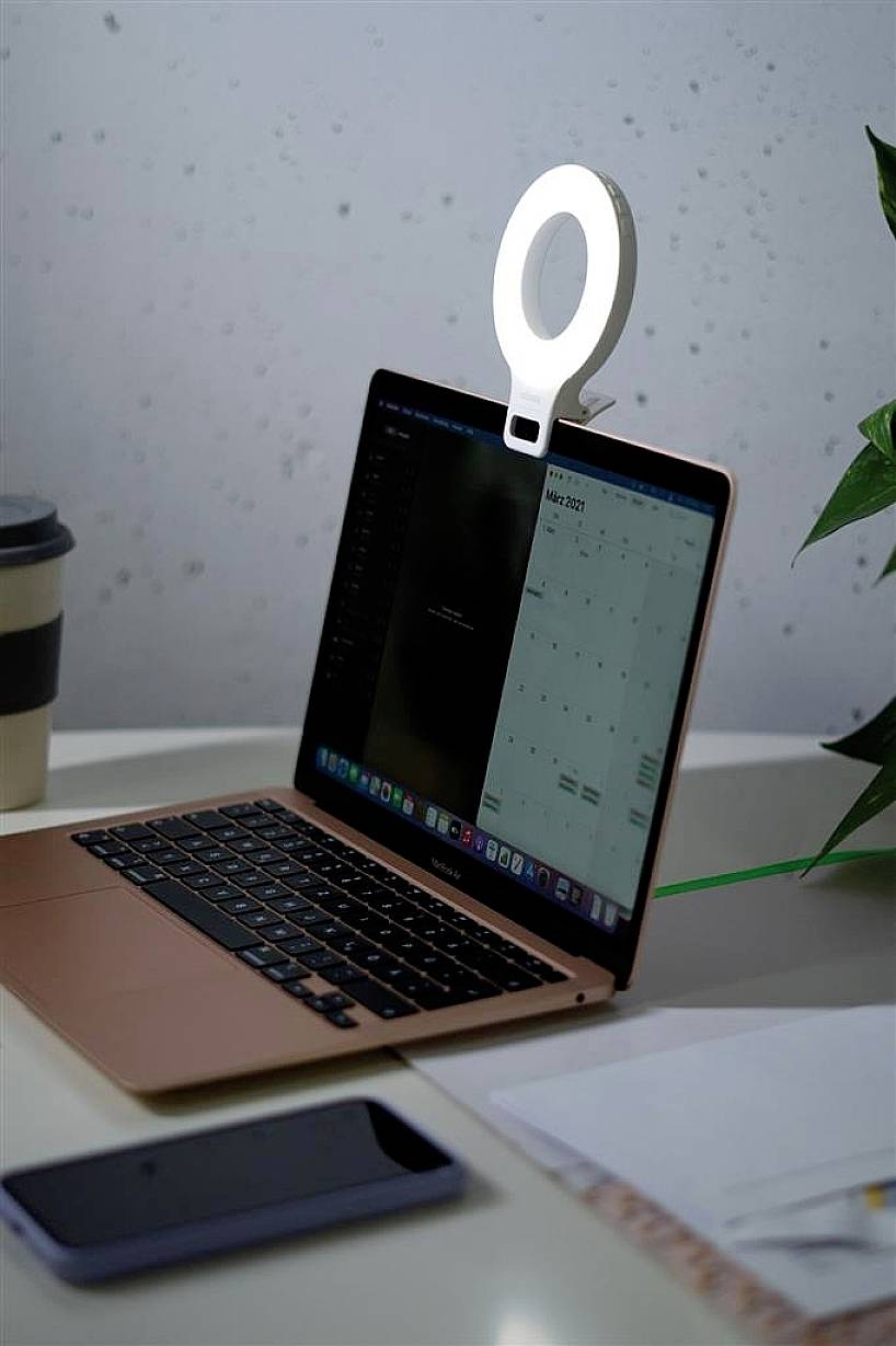 Un laptop su una scrivania con un supporto luminoso rotondo, accanto a un bicchiere di caffè, uno smartphone e delle piante. Sullo schermo è visibile un calendario.