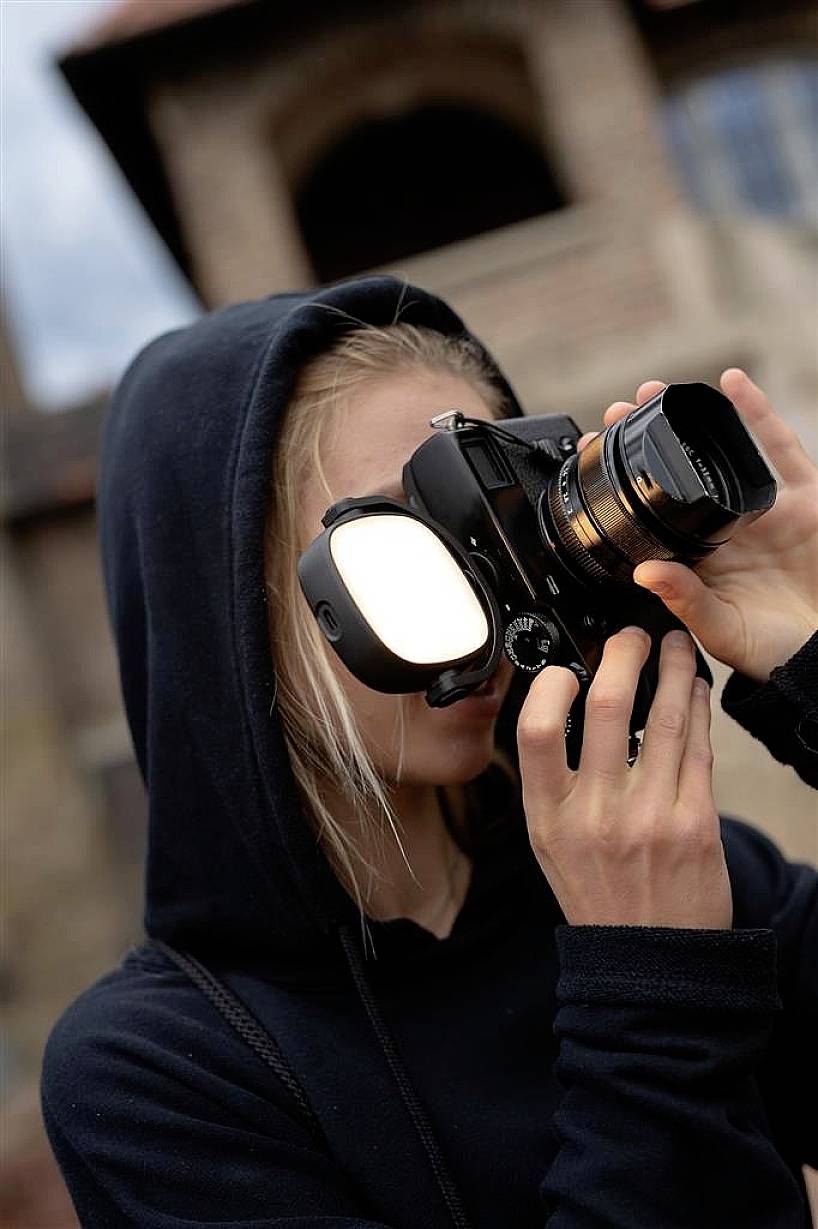 Una persona con un felpa nera sta fotografando con una macchina fotografica. Lo sfondo è sfocato e mostra un edificio.
