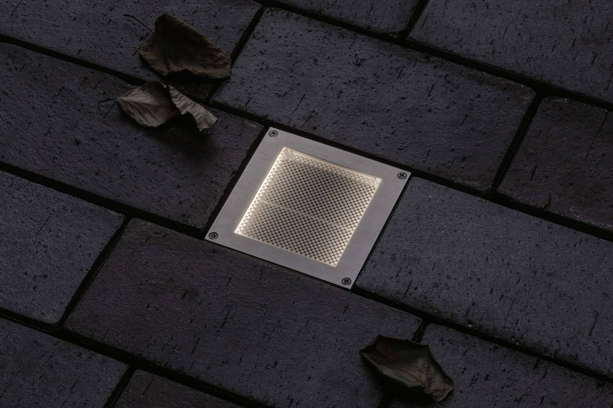 Faretto LED da incasso a pavimento illuminato, integrato nei mattoni per pavimentazione e circondato da foglie secche.