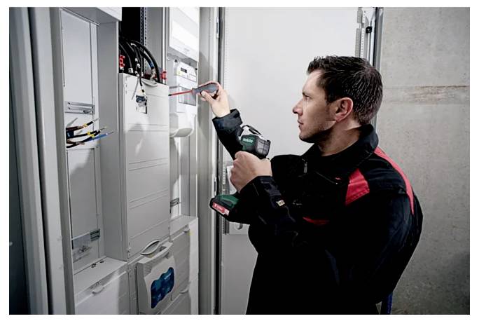 Un tecnico in divisa usa un cacciavite e un trapano per lavorare su pannelli elettrici in una sala di controllo.