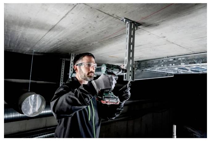 Una persona che indossa occhiali protettivi e guanti utilizza un trapano elettrico per installare supporti per soffitti in un ambiente industriale.