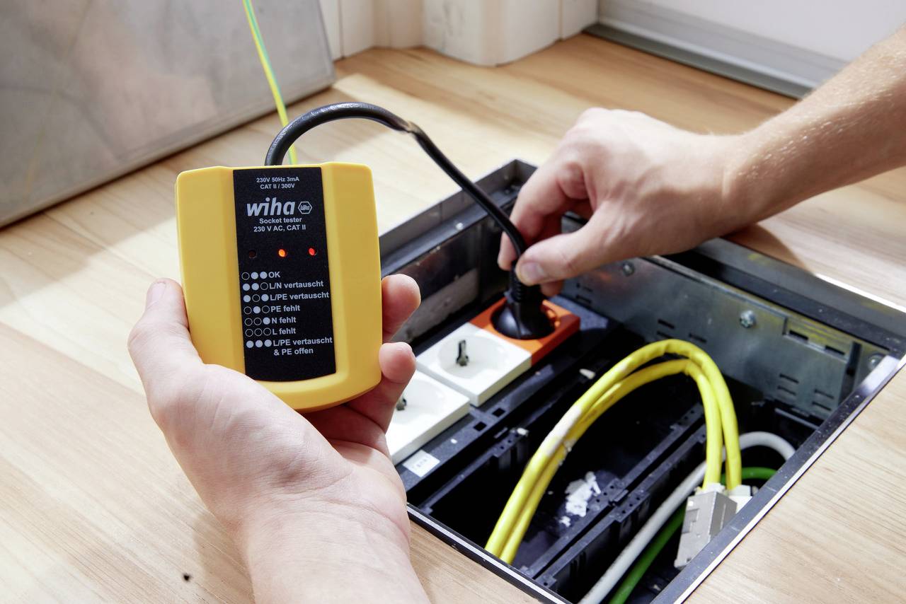 Una persona sta utilizzando un dispositivo elettrico di prova per verificare i cavi in un canale di pavimentazione aperto. Una luce rossa indica lo stato del test.