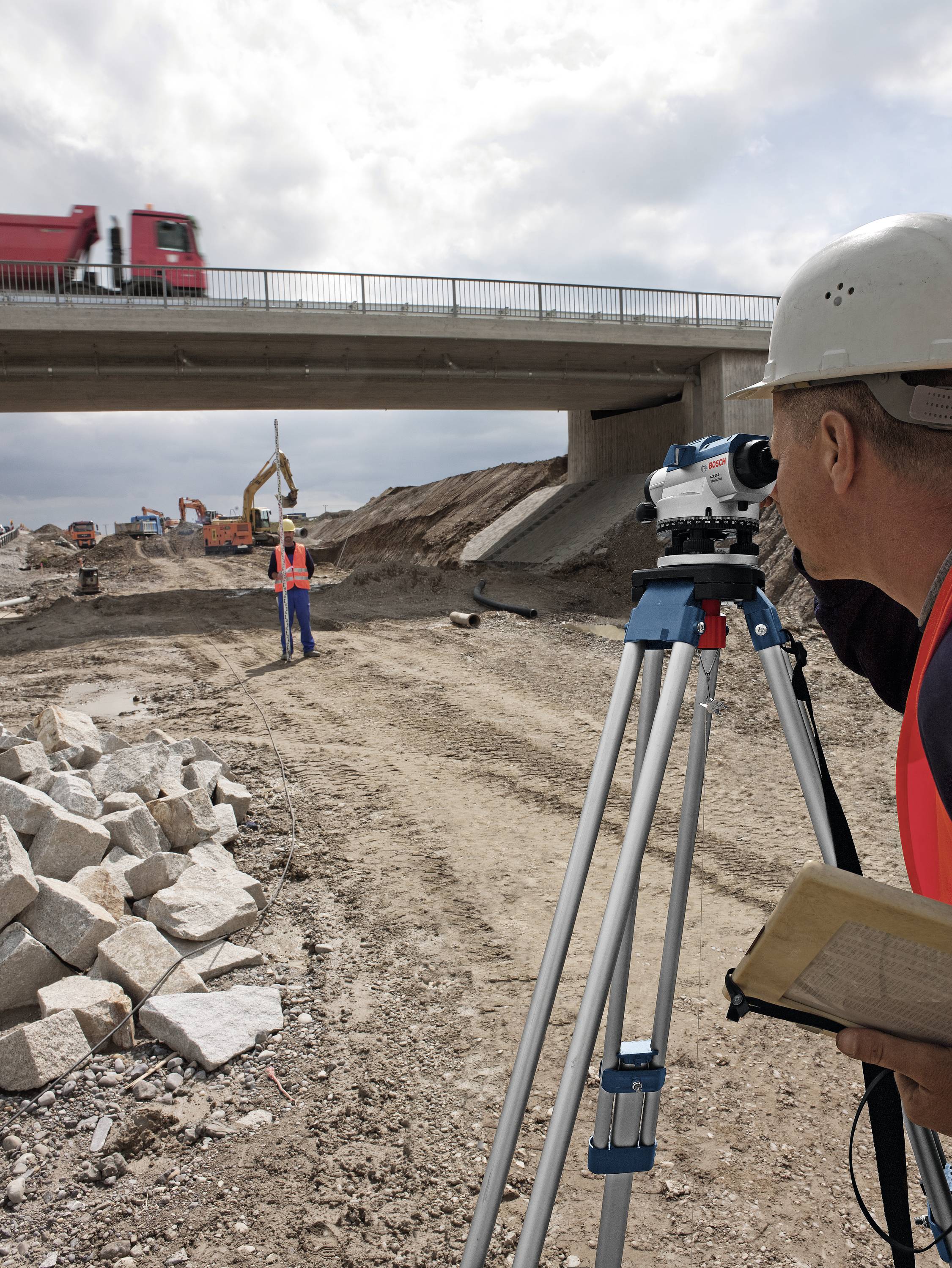Ingegnere topografo con casco e strumento di misurazione osserva i lavori di costruzione sotto un ponte, mentre un veicolo passa accanto. Sullo sfondo, escavatori e operai.