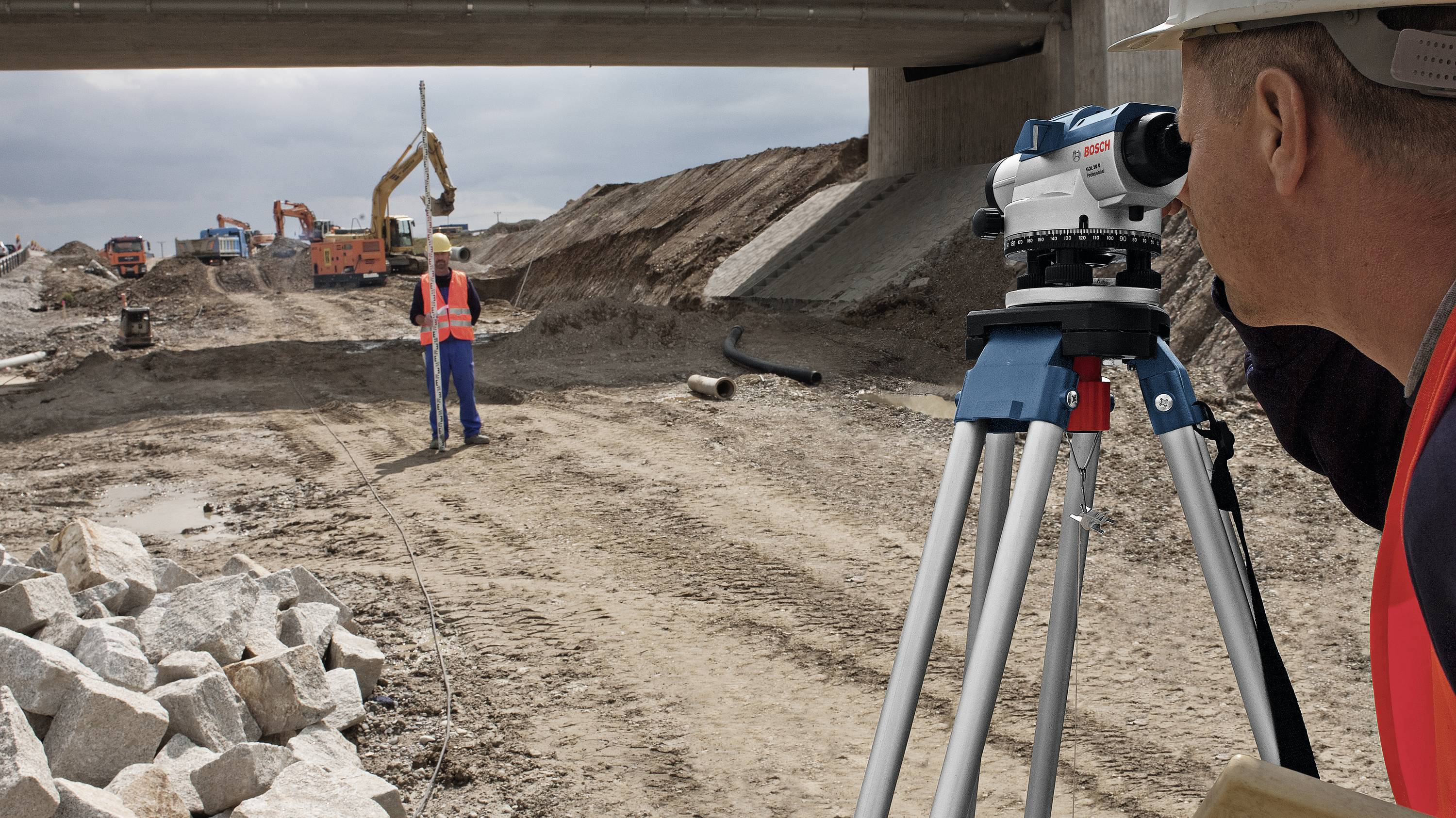 Un lavoratore edile utilizza uno strumento di misurazione per rilevare un cantiere sotto un ponte. Sullo sfondo sono visibili degli escavatori.