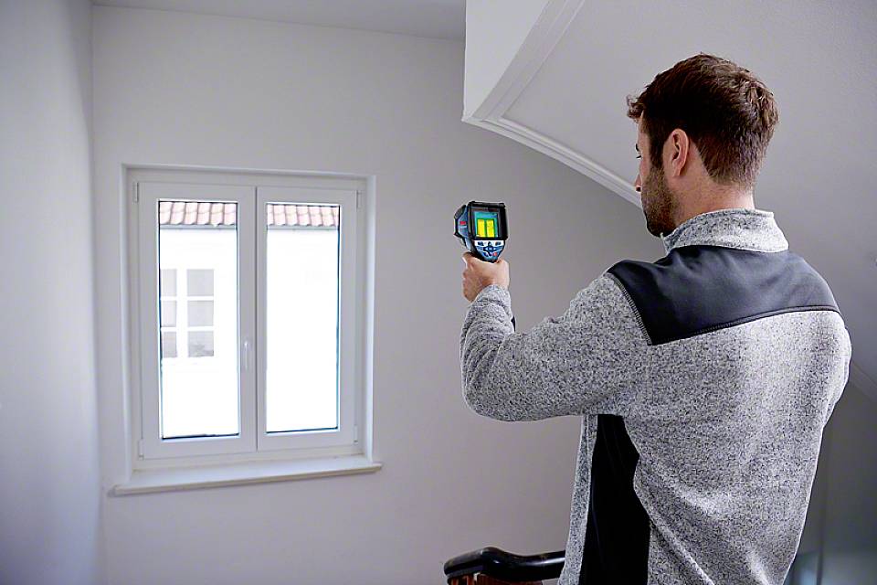 Un uomo utilizza una termocamera per verificare l'isolamento di una finestra in una stanza bianca.