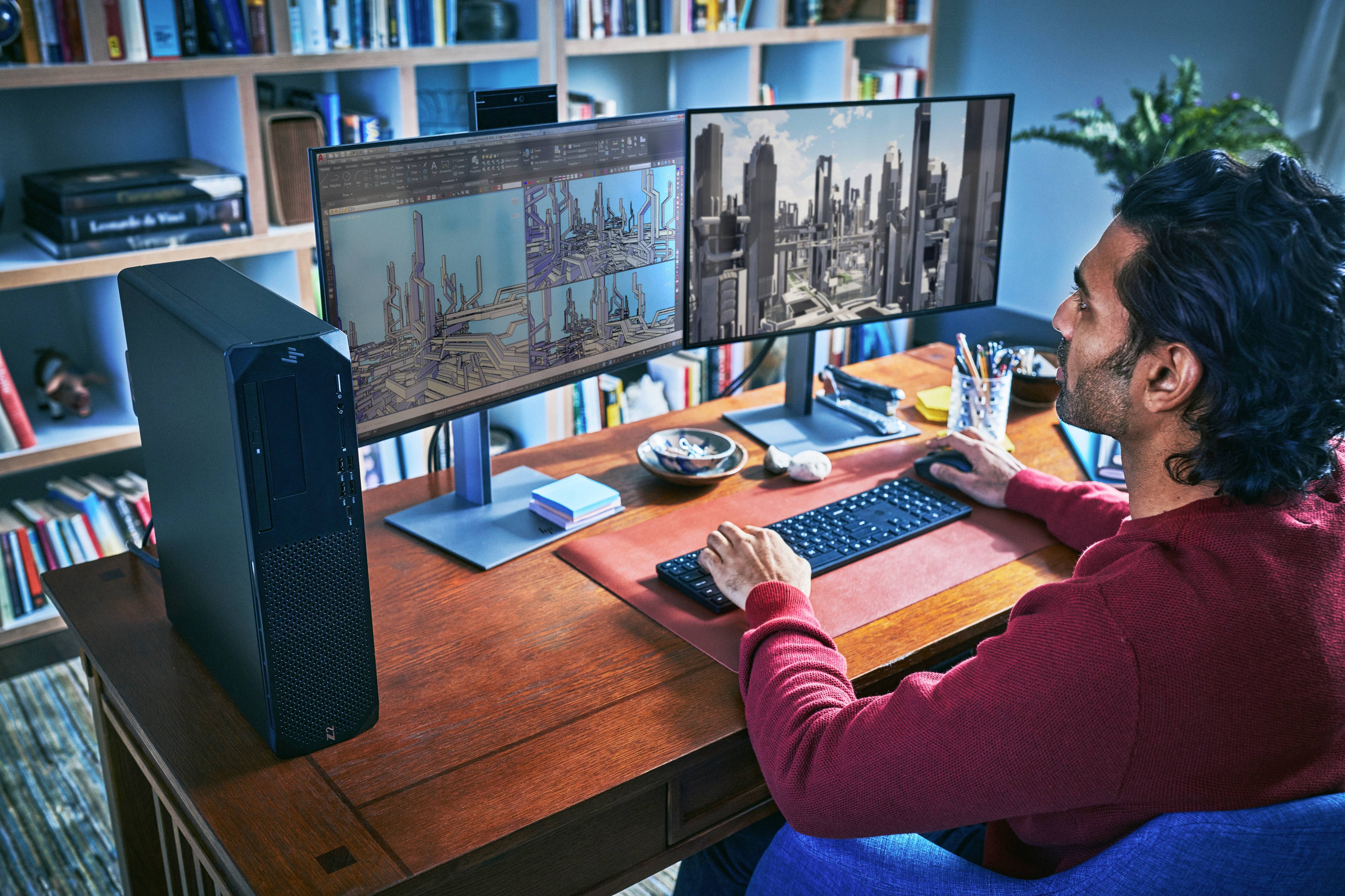 Un uomo lavora a una scrivania con due monitor che mostrano grafici complessi e modelli 3D. Sullo sfondo, uno scaffale pieno di libri.