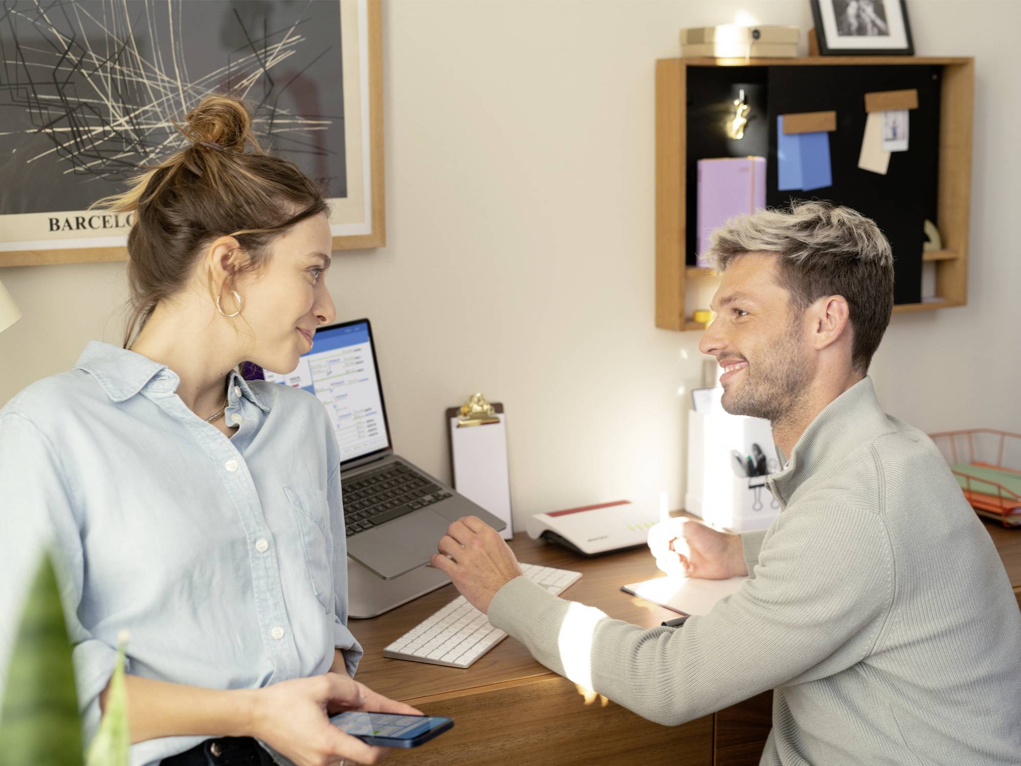 Un uomo e una donna conversano a una scrivania con un laptop. Ambiente d'ufficio con documenti e decorazioni sullo sfondo.