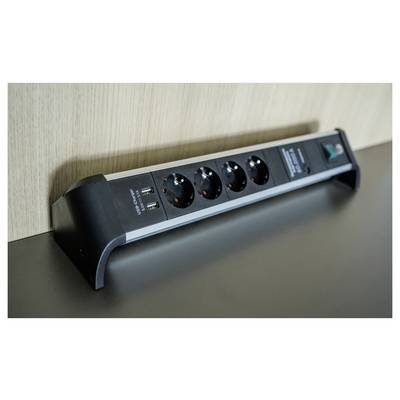 Ciabatta di alimentazione nera con quattro prese di corrente e due porte USB montata lungo una parete su una scrivania o uno scaffale.