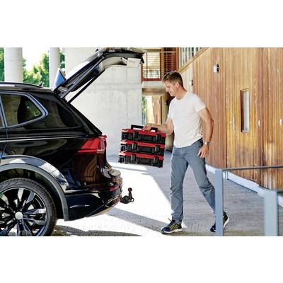 Un uomo in abiti casual sta caricando diverse cassette degli attrezzi rosse e nere nel bagagliaio di un SUV nero davanti a un edificio moderno.