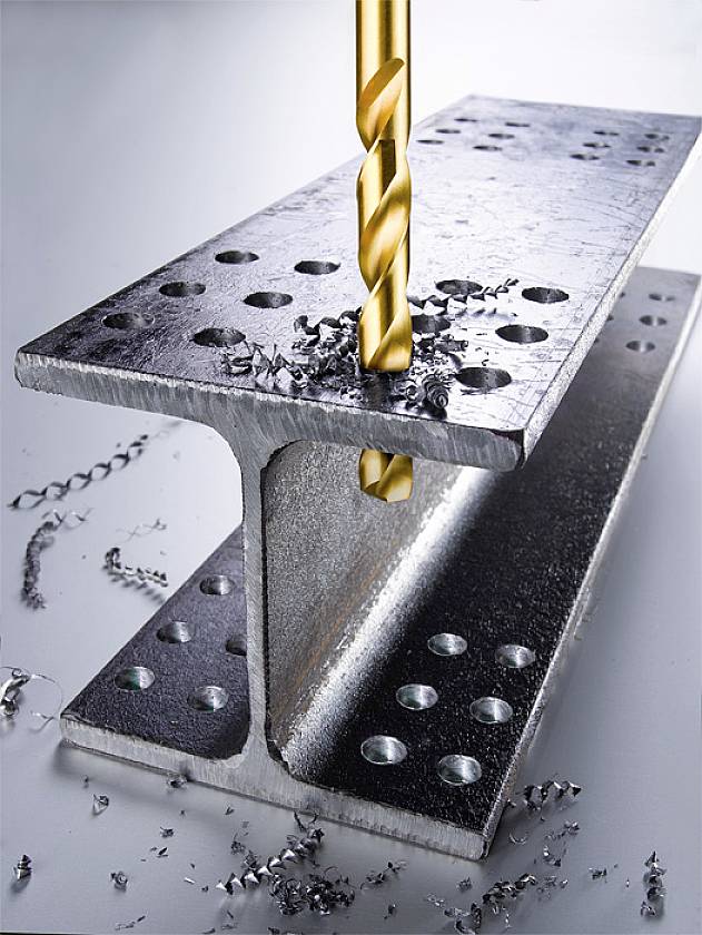 Un trapano dorato perfora un profilo portante in metallo con diversi fori, trucioli metallici sono sparsi intorno al profilo.
