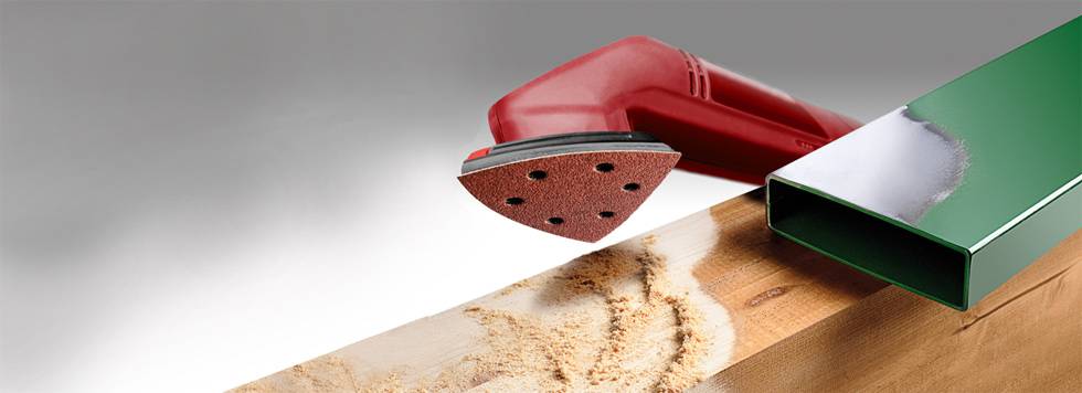 Un piccolo dispositivo di levigatura rosso sta levigando una superficie in legno; accanto ad esso giace un tubo metallico lucido di colore verde.
