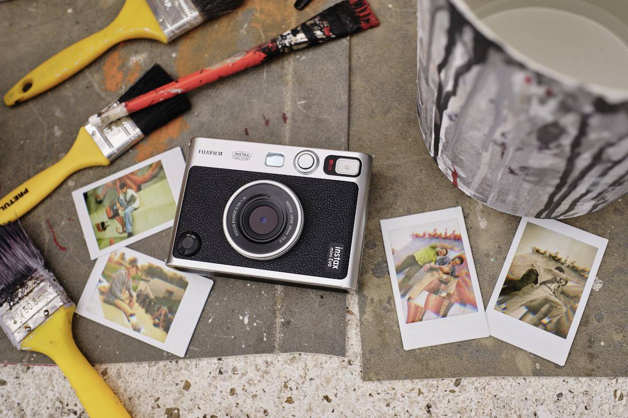 Una fotocamera è posizionata su un tavolo accanto a pennelli e tre foto stampate. Nel quadro è visibile anche un secchio dipinto.