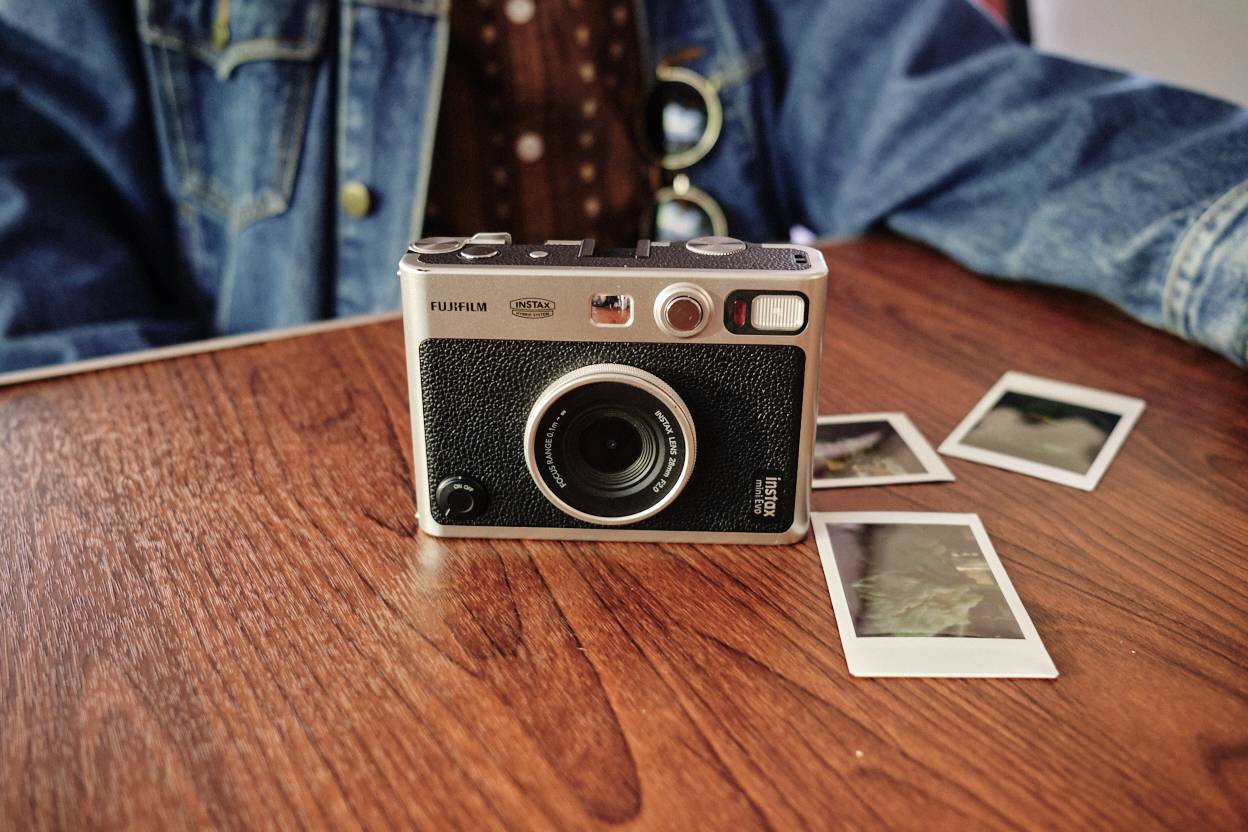 Una macchina fotografica analogica è posizionata su un tavolo di legno, circondata da tre istantanee. Una persona indossa una giacca di jeans sullo sfondo.