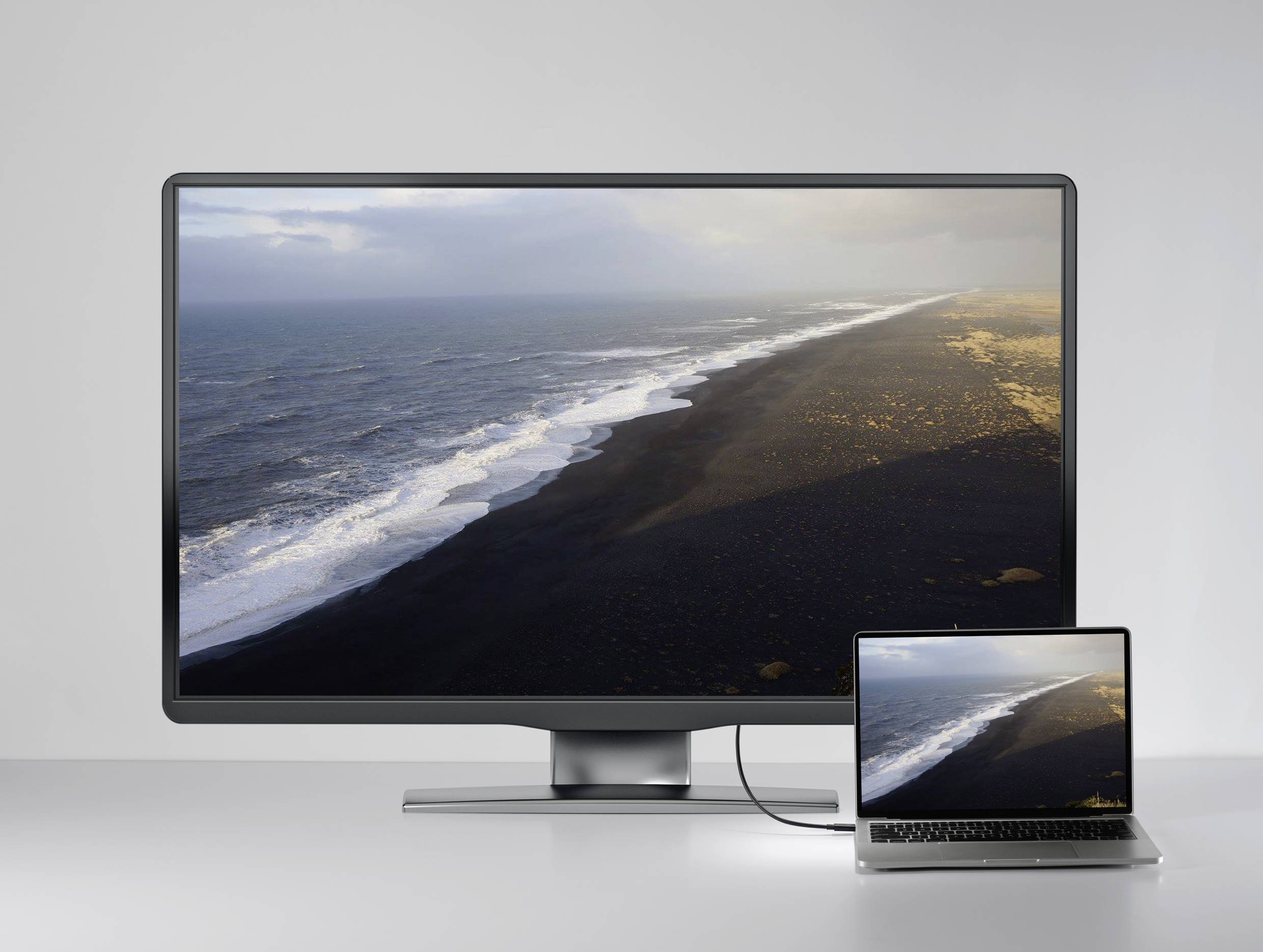Un grande monitor e un laptop collegato mostrano la stessa immagine di una linea costiera con sabbia nera e onde.