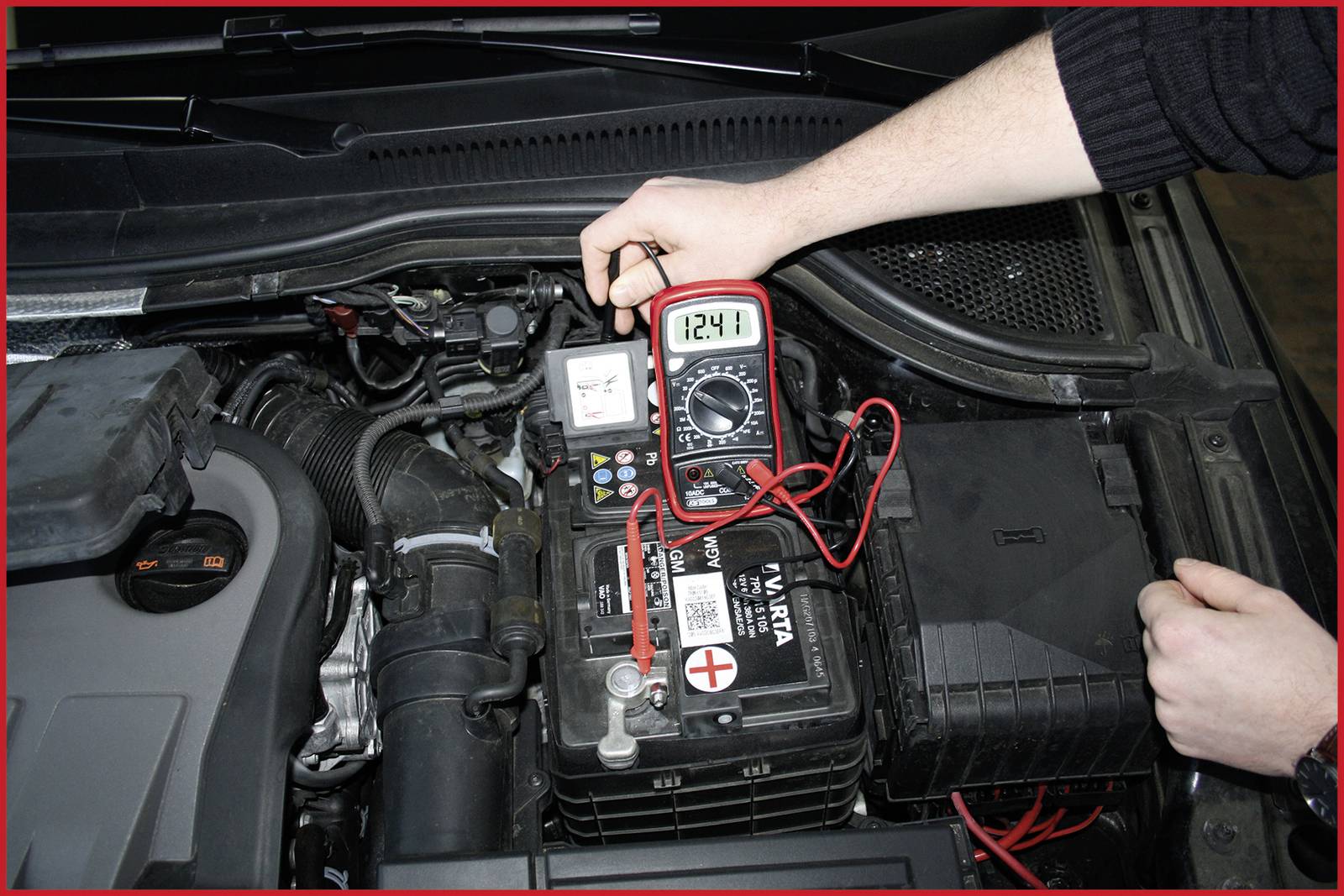 Una persona misura la tensione di una batteria per auto con un multimetro. Il display mostra '12,41'.