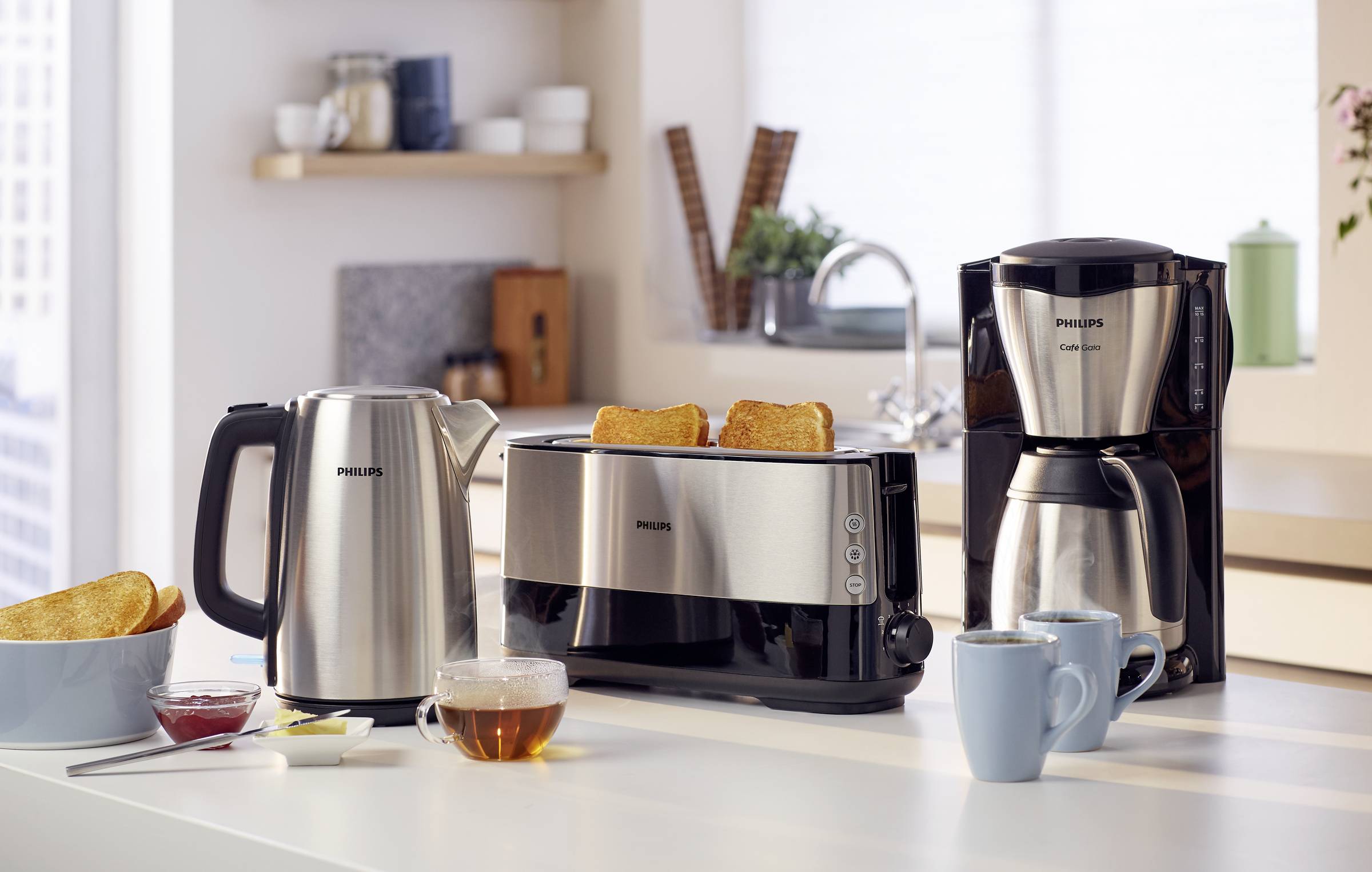 'Set di elettrodomestici su piano di lavoro: bollitore, tostapane a due fette, macchina per caffè a filtro; sullo sfondo scaffali.'