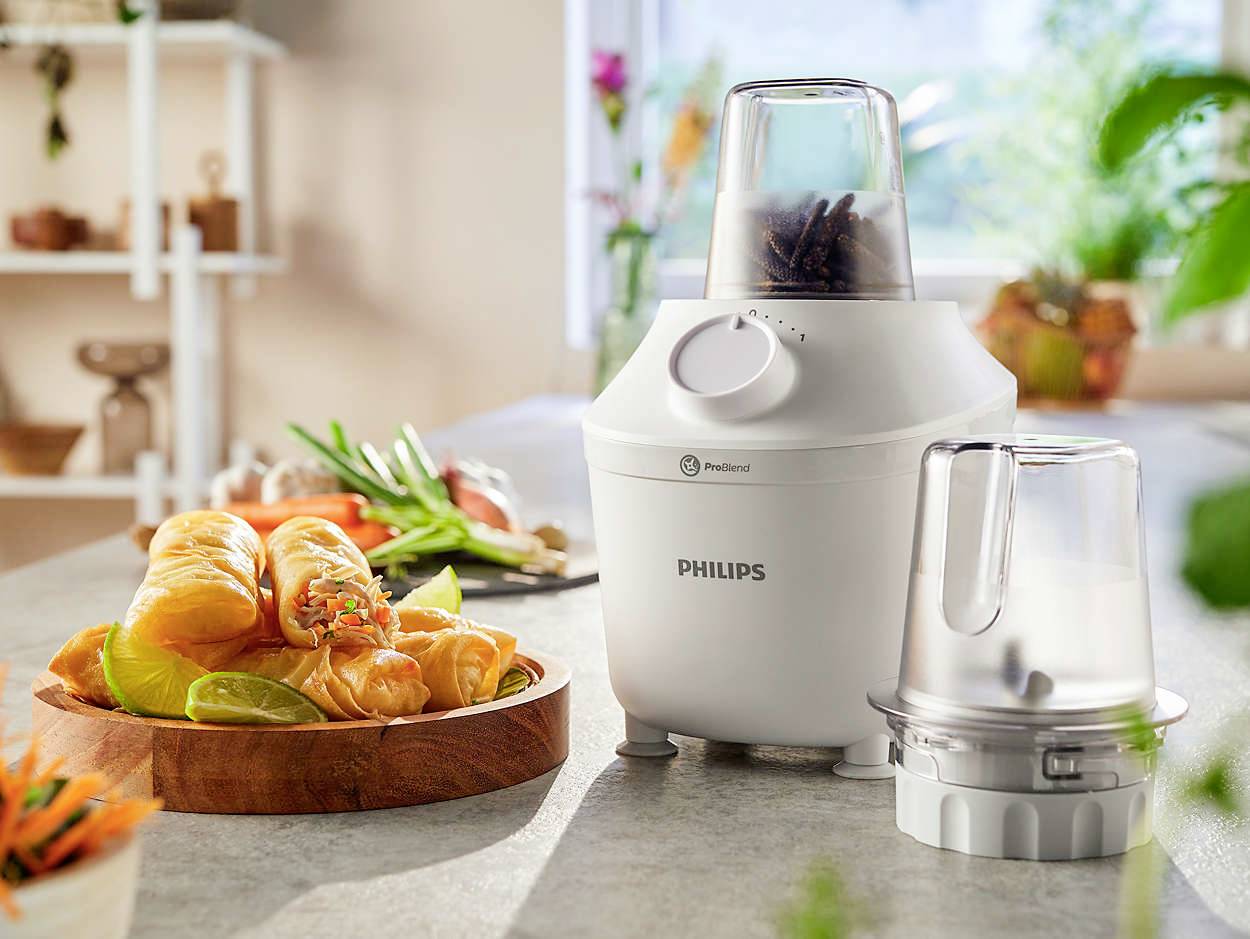 Un mixer bianco Philips è posizionato su un tavolo da cucina accanto a un piatto con involtini primavera. Sullo sfondo si intravedono erbe aromatiche da cucina.