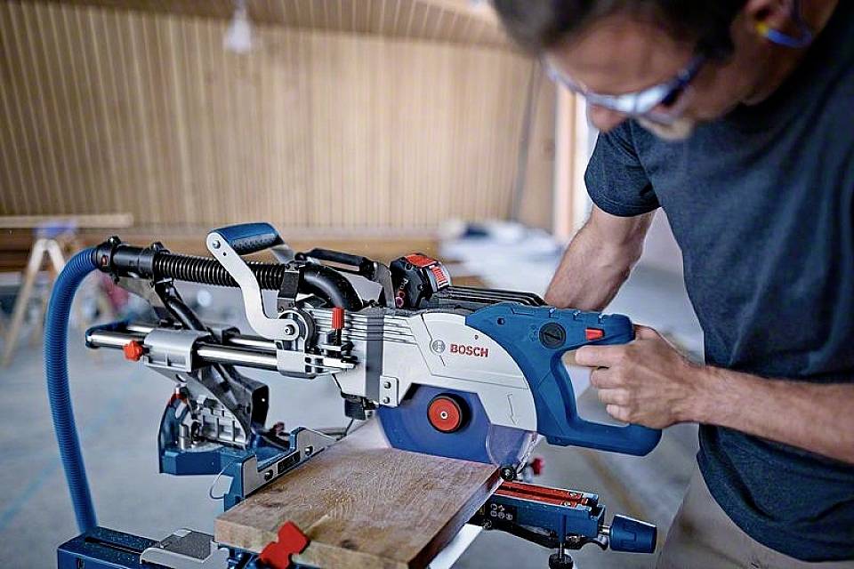 Un uomo in officina usa una sega da taglio elettrica per tagliare una tavola di legno. L'ambiente appare artigianale e tecnico.