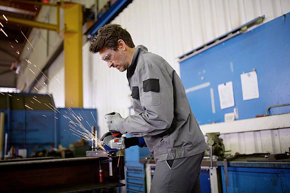Un uomo in abbigliamento protettivo sta levigando il metallo in un laboratorio. Scintille volano dalla smerigliatrice. Sullo sfondo si vedono attrezzature da officina.