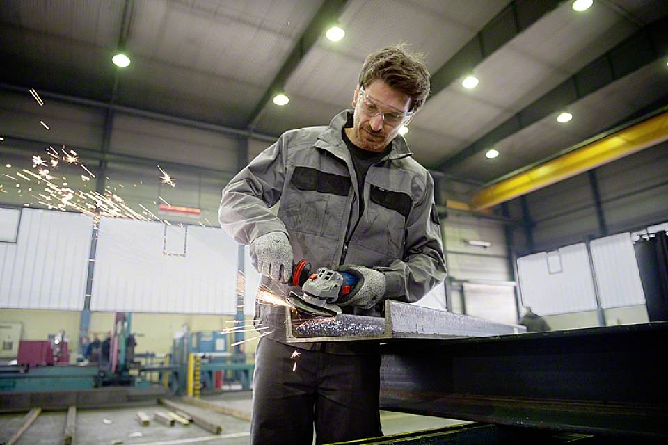 Un uomo sta lavorando un pezzo di metallo con un angolare in un capannone industriale. Scintille volano mentre lavora con concentrazione.
