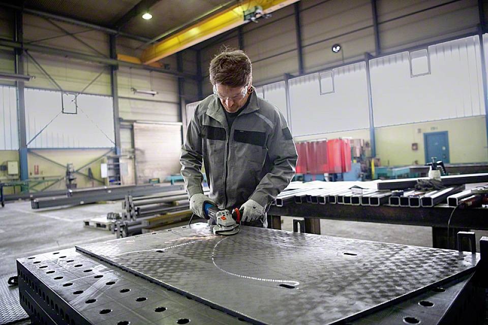 Un uomo in abiti da lavoro sta lavorando una lastra di metallo con un utensile da taglio in un grande capannone industriale.