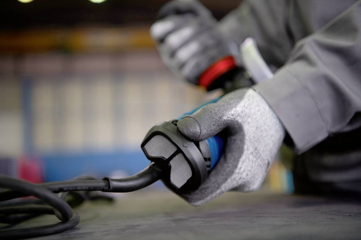 Una persona con guanti grigi tiene la spina di un cavo elettrico, presumibilmente in un ambiente di officina.