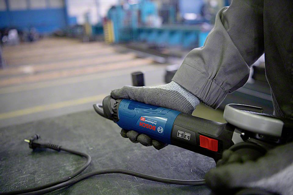 Una persona tiene un smerigliatrice angolare blu in un laboratorio. Un cavo elettrico è allentato sul tavolo davanti.