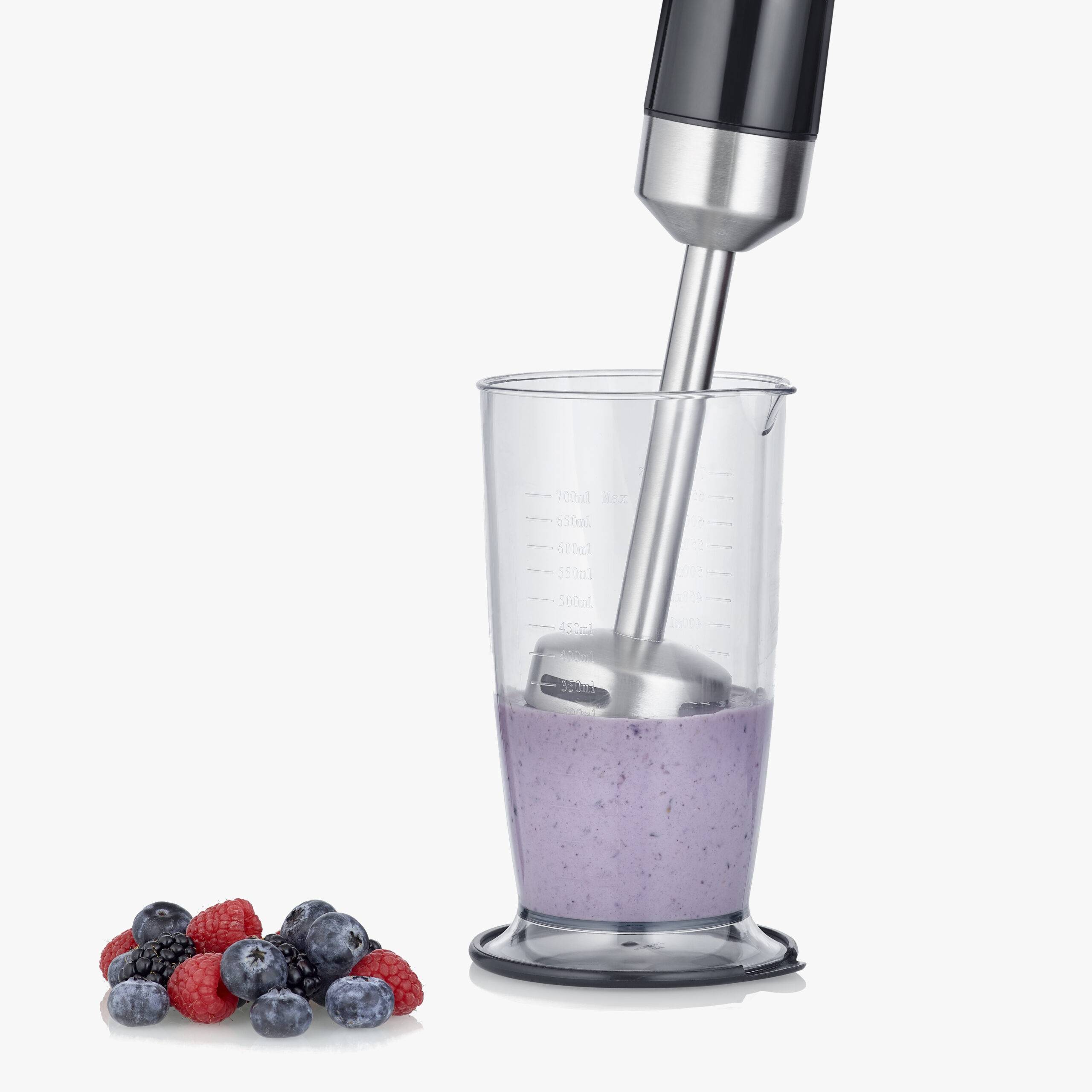Un frullatore sta frullando un smoothie viola in un misurino. Accanto ci sono alcuni lamponi e mirtilli su un tavolo.