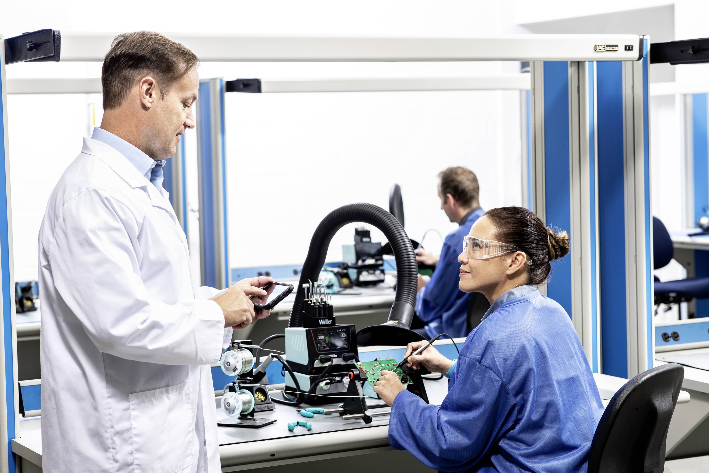 Un uomo e una donna in camici da lavoro stanno lavorando su dispositivi elettronici in un laboratorio. Si comunicano a vicenda.