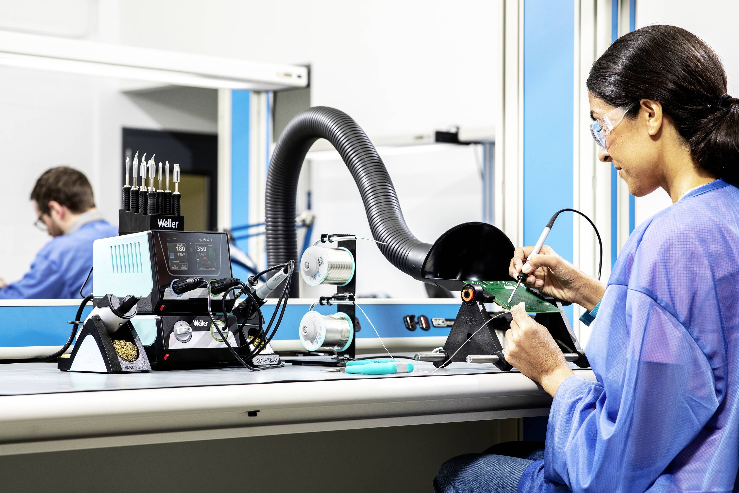 Una donna lavora con un saldatore su una scheda elettronica in un laboratorio, con un sistema di aspirazione e utensili sullo sfondo.