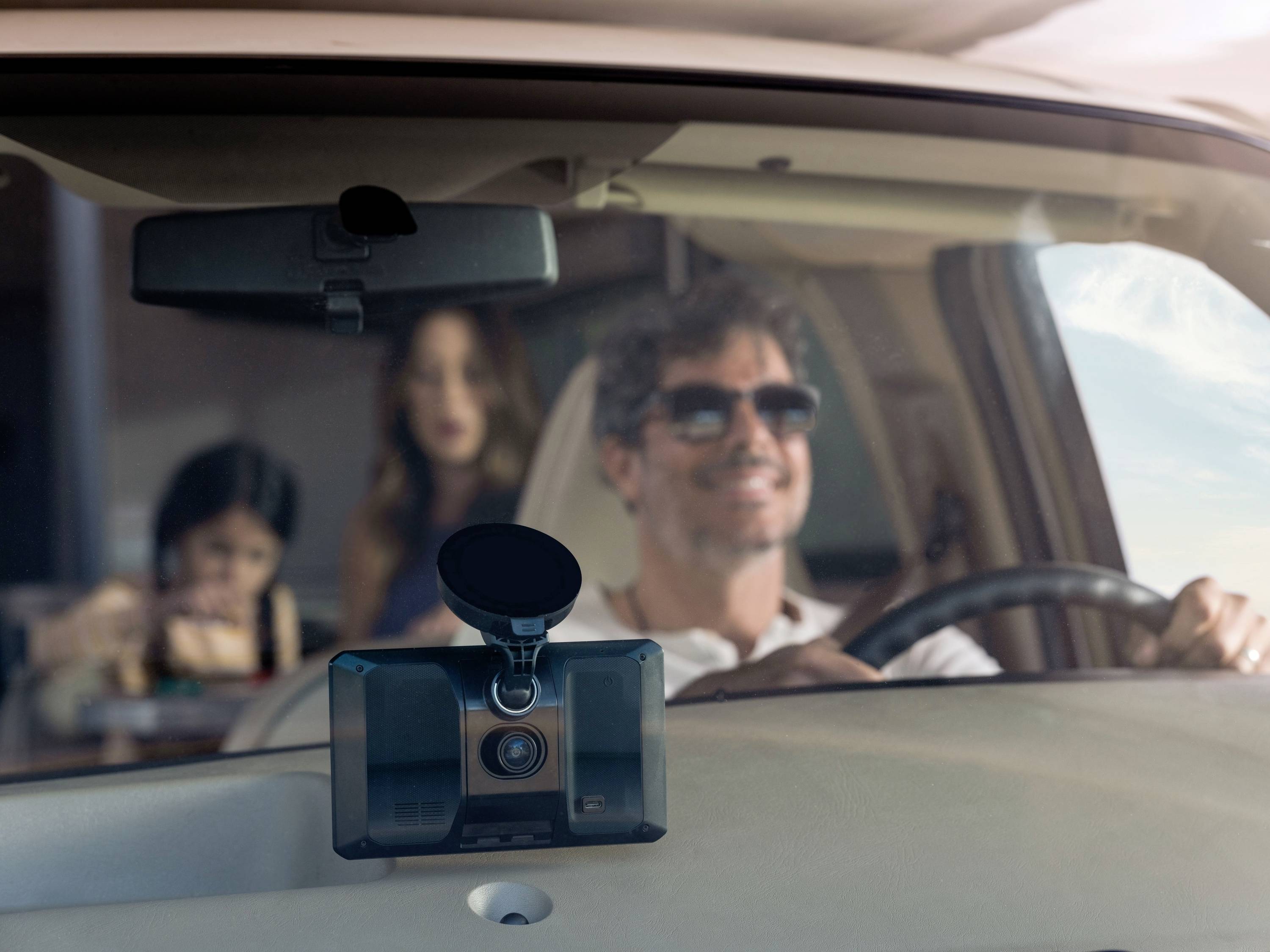 Un uomo con gli occhiali da sole guida un'auto. Una donna e un bambino sono visibili sullo sfondo. Si tratta di un interno auto moderno.