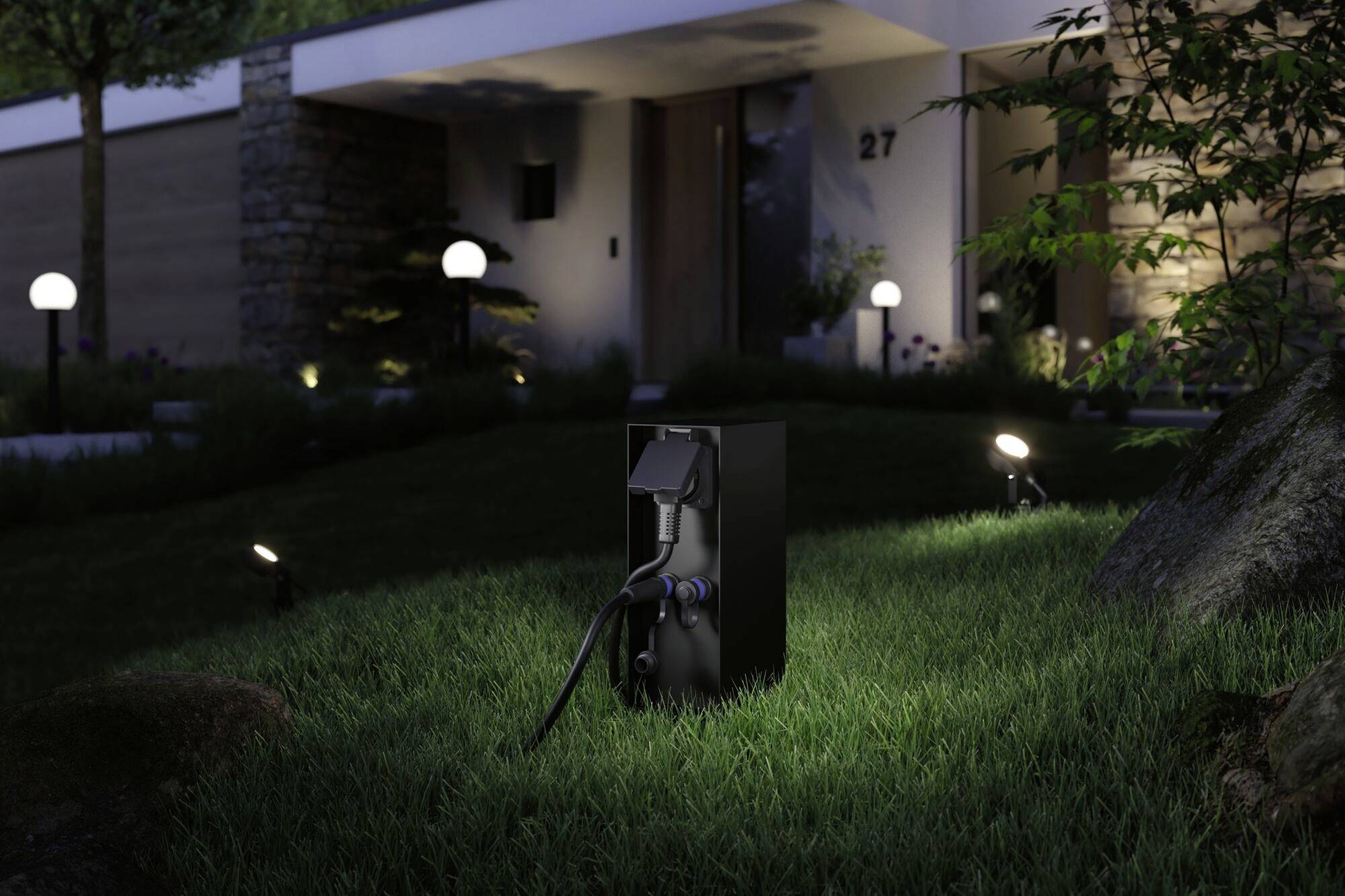 Esterno di una casa di notte con un giardino illuminato. In primo piano, una presa elettrica nera con un cavo collegato.
