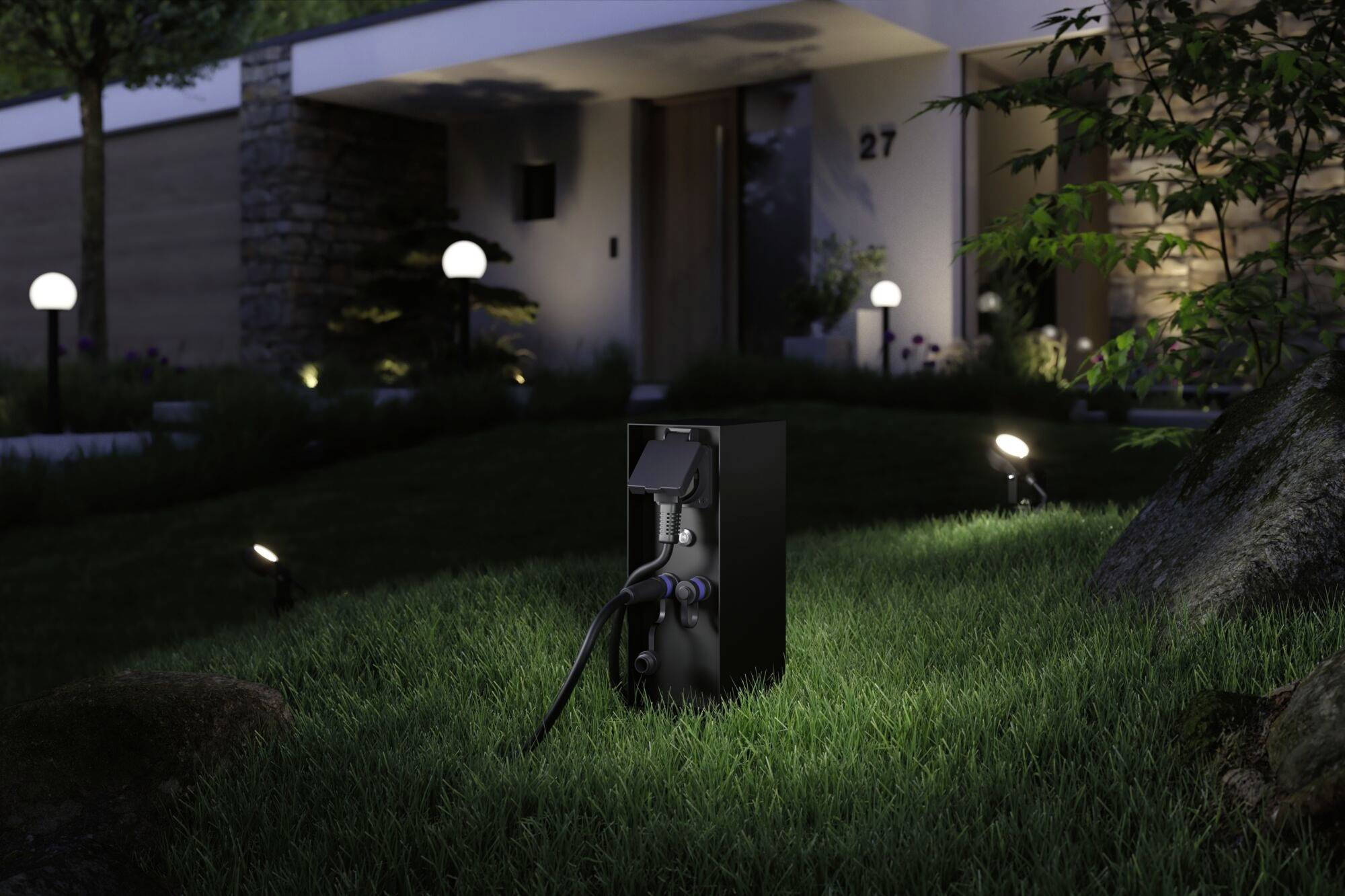 Una stazione di ricarica nera per auto elettriche è posizionata su un prato davanti a una casa moderna di notte, illuminata da piccole luci a terra.