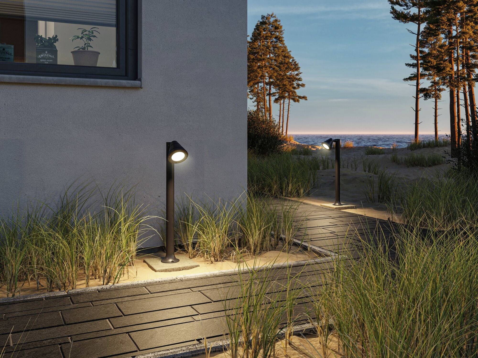Un sentiero illuminato da lanterne a terra conduce a una spiaggia al tramonto. A sinistra si trova un edificio. Alte erbe costeggiano il percorso.