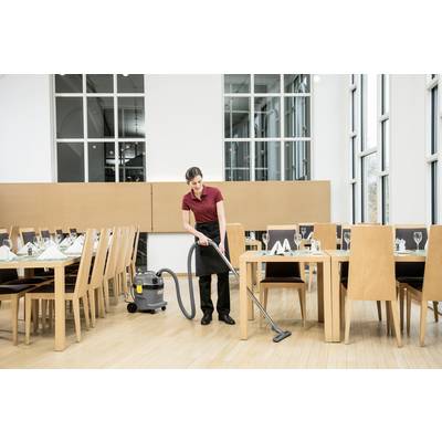 Una persona aspira il pavimento in un ristorante vuoto con un aspirapolvere da tavolo. I tavoli sono apparecchiati, con grandi finestre sullo sfondo.