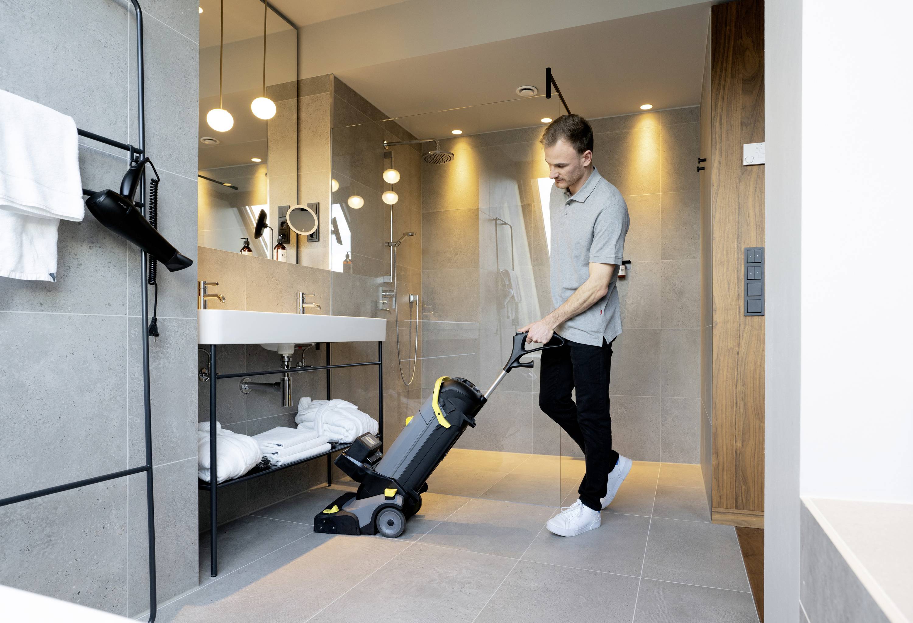Un uomo sta utilizzando una macchina per la pulizia del pavimento in un bagno moderno con doccia, grandi specchi e asciugamani sullo scaffale.