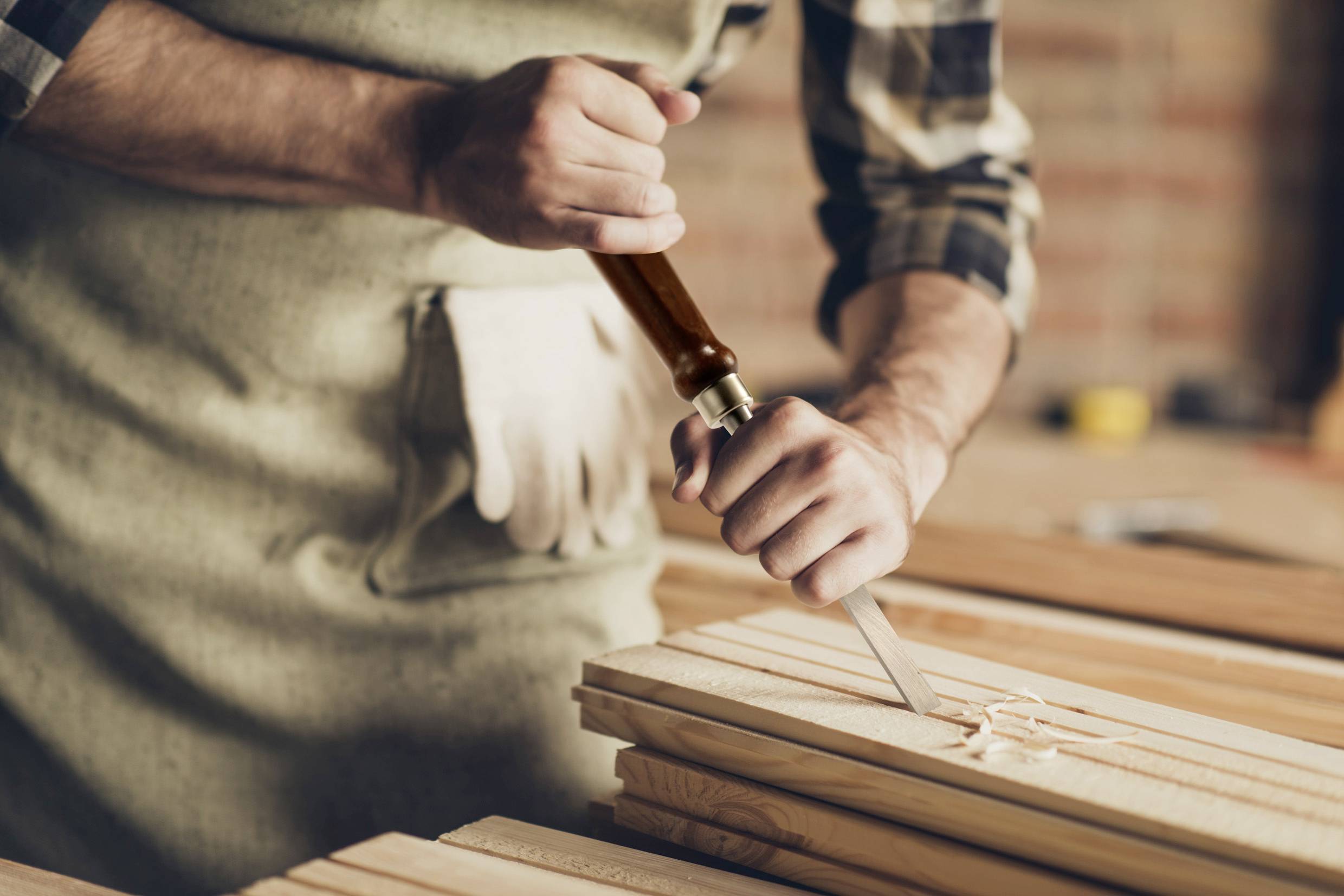 Una persona con un grembiule incide un motivo su un pezzo di legno con uno scalpello. Le mani e lo strumento sono a fuoco.