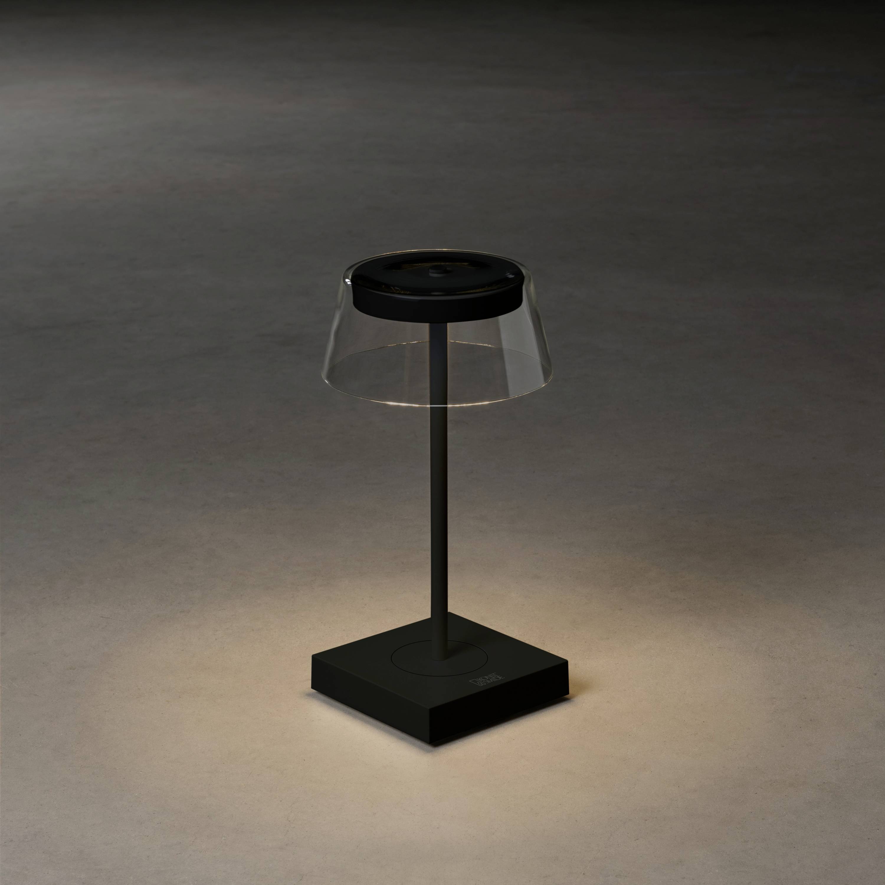 Una moderna lampada da tavolo con base nera e paralume trasparente, illumina un'area in uno spazio dall'arredamento minimalista.