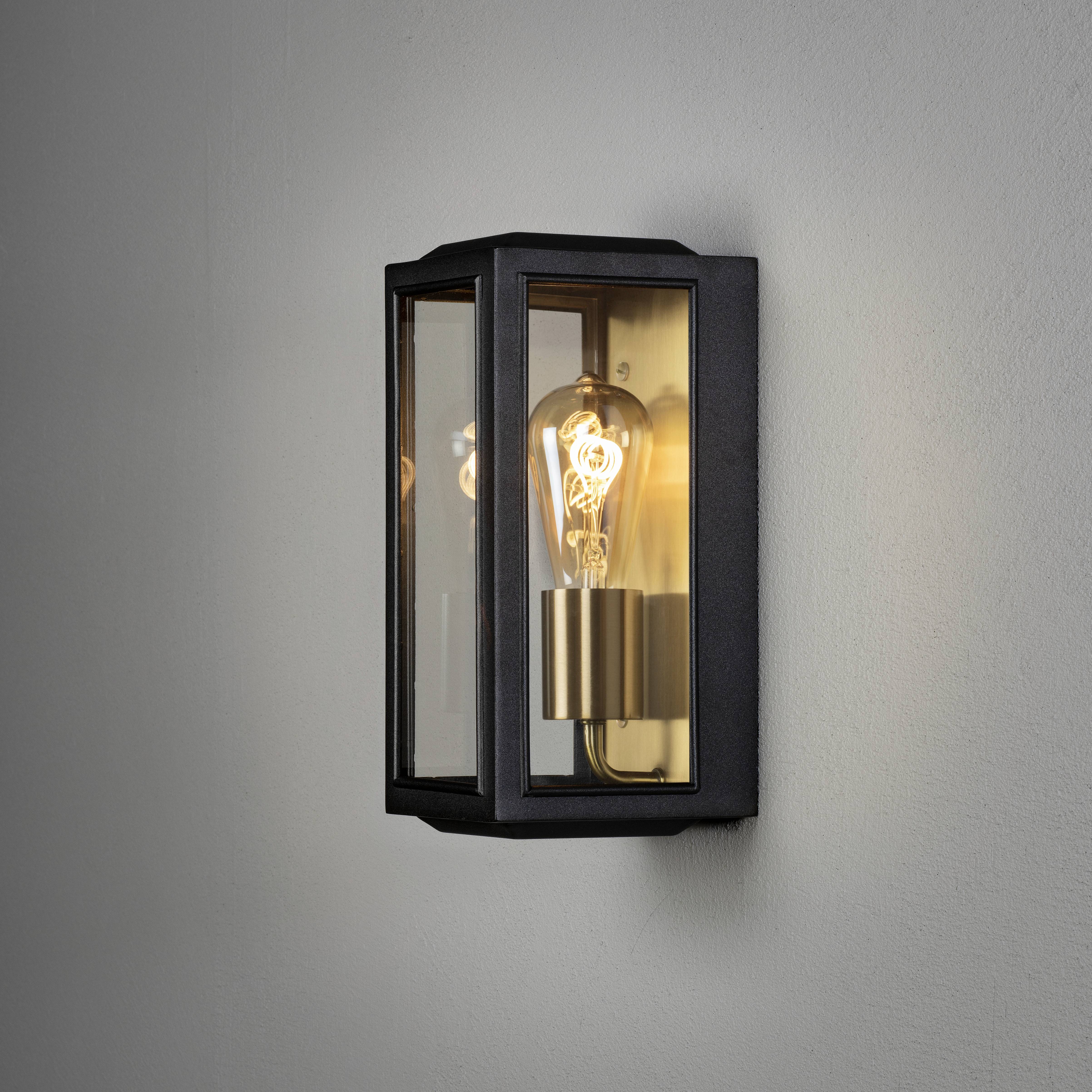 'Applique con cornice in metallo nero, pannelli di vetro trasparenti e lampadina a vista, accesa su una parete grigia.'