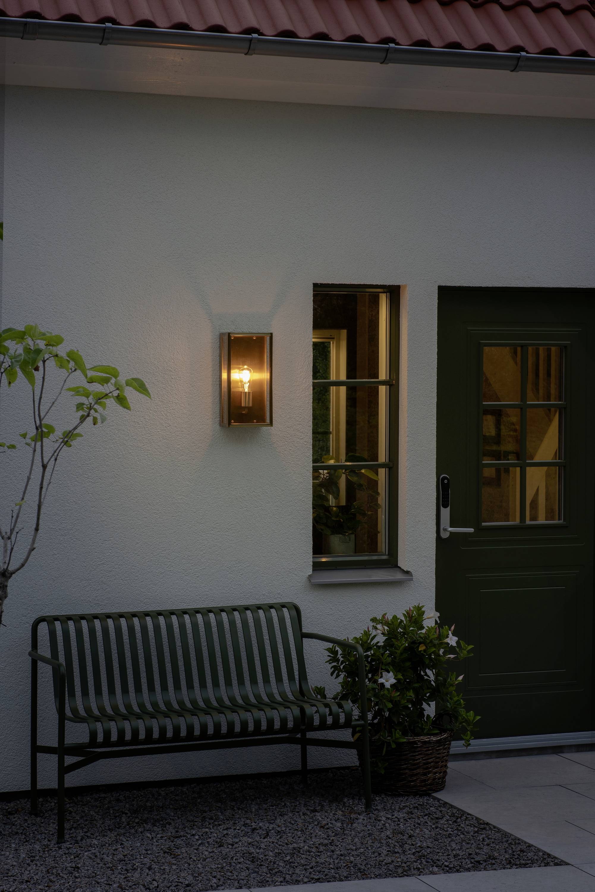 Un'illuminazione esterna illumina un muro esterno con una finestra e una porta verde. Sotto, è posizionata una panchina verde accanto a un cesto di piante.