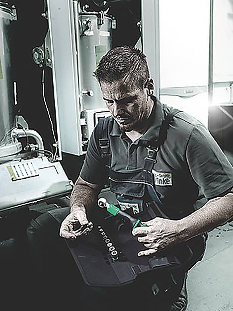 Un tecnico in tuta da lavoro, inginocchiato, esamina dei componenti con gli strumenti vicini, probabilmente intento a eseguire interventi di manutenzione su macchinari industriali.