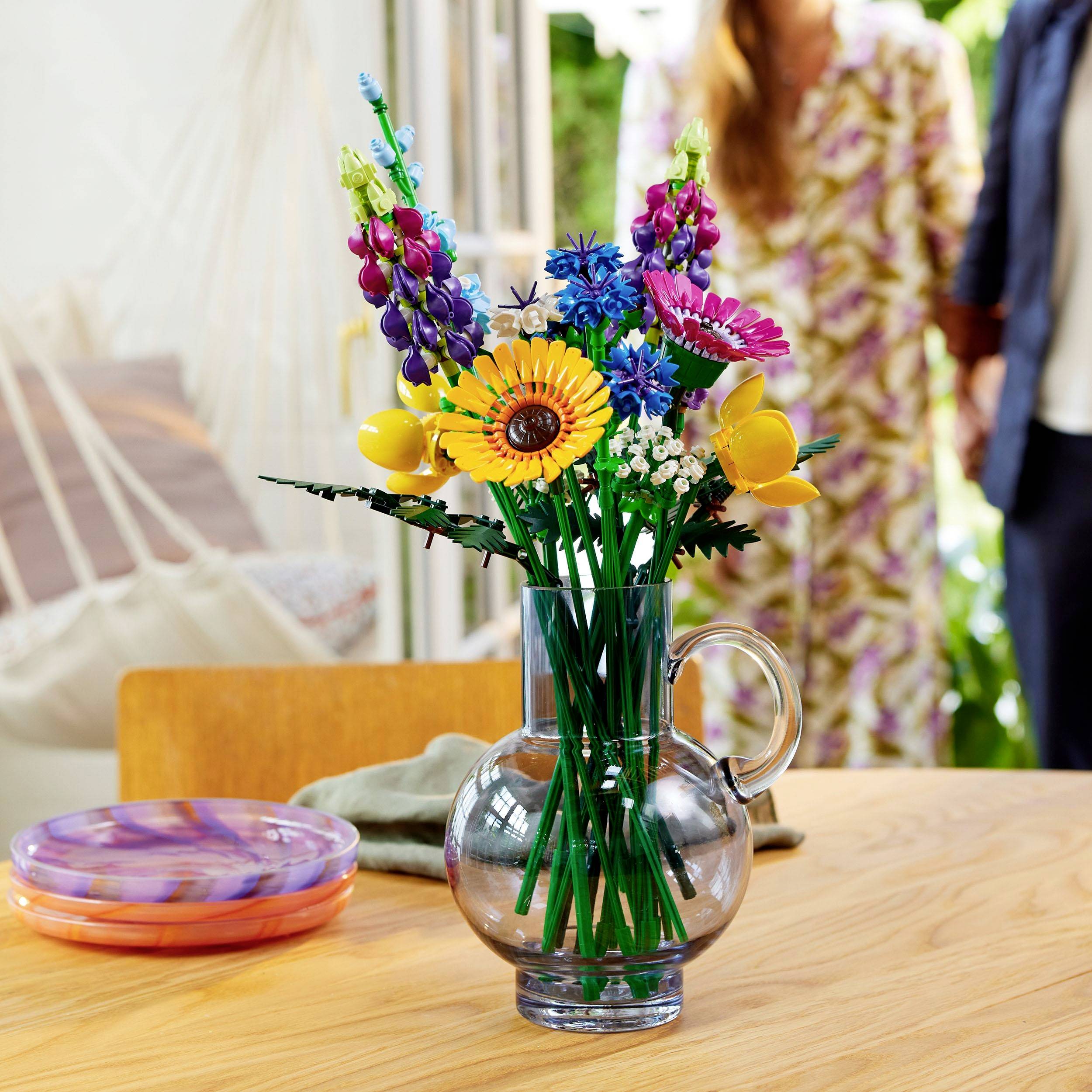 Un mazzo colorato di fiori misti in un vaso trasparente è poggiato su un tavolo di legno, sullo sfondo si vedono due persone e una finestra aperta.