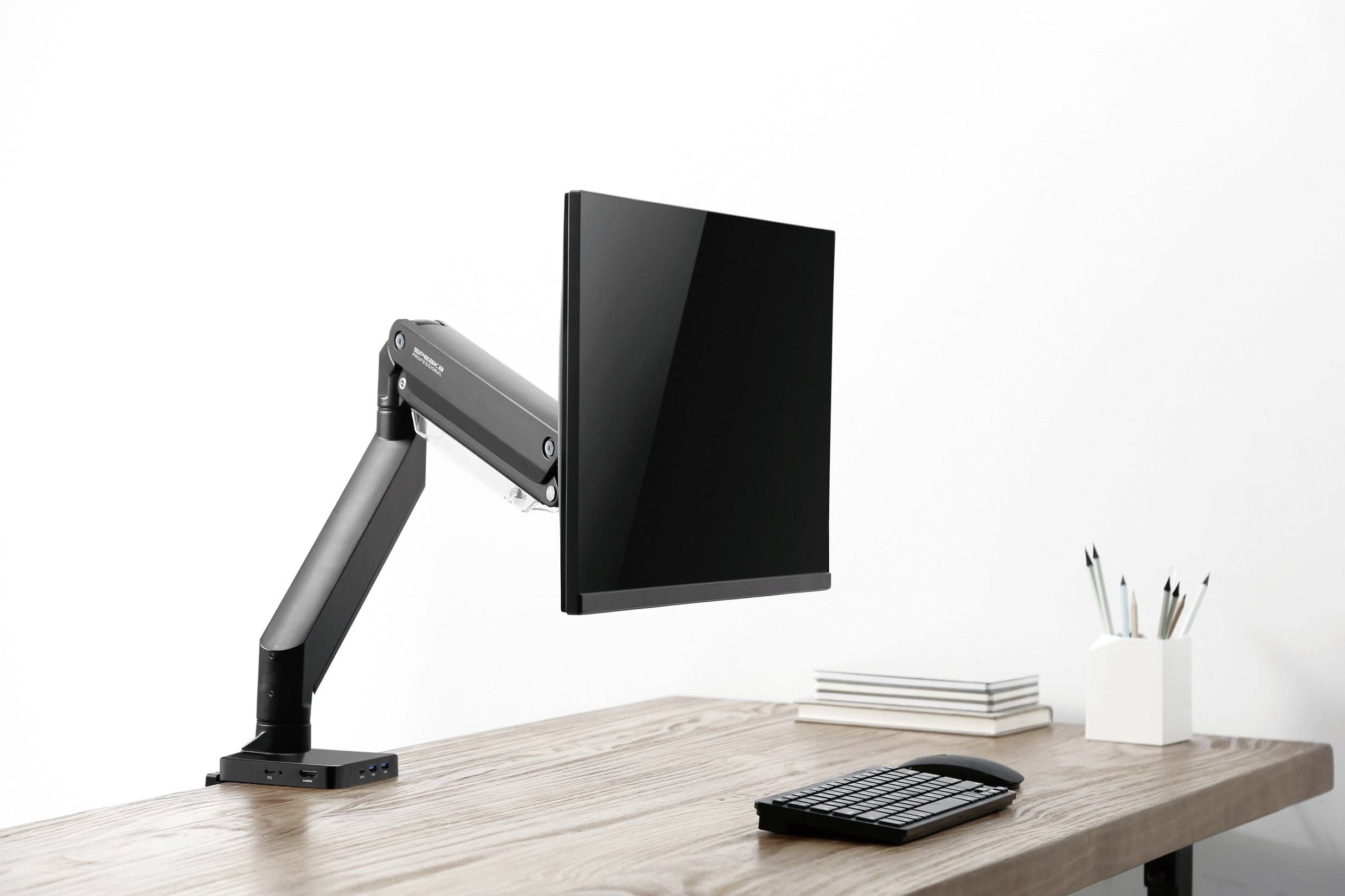 Un monitor è montato su un braccio flessibile sopra una scrivania. Sul tavolo ci sono quaderni e penne.