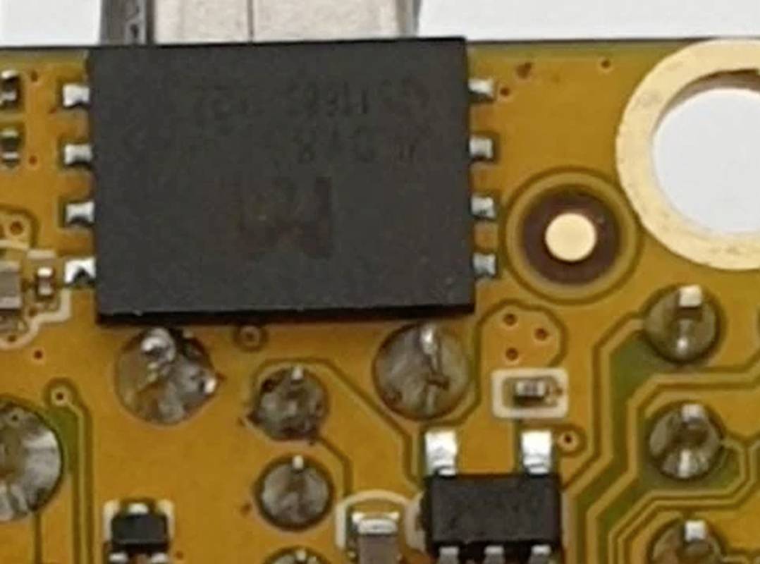 Primo piano di un circuito stampato con vari componenti elettronici. Include un chip nero, contatti di saldatura e piste conduttrici.