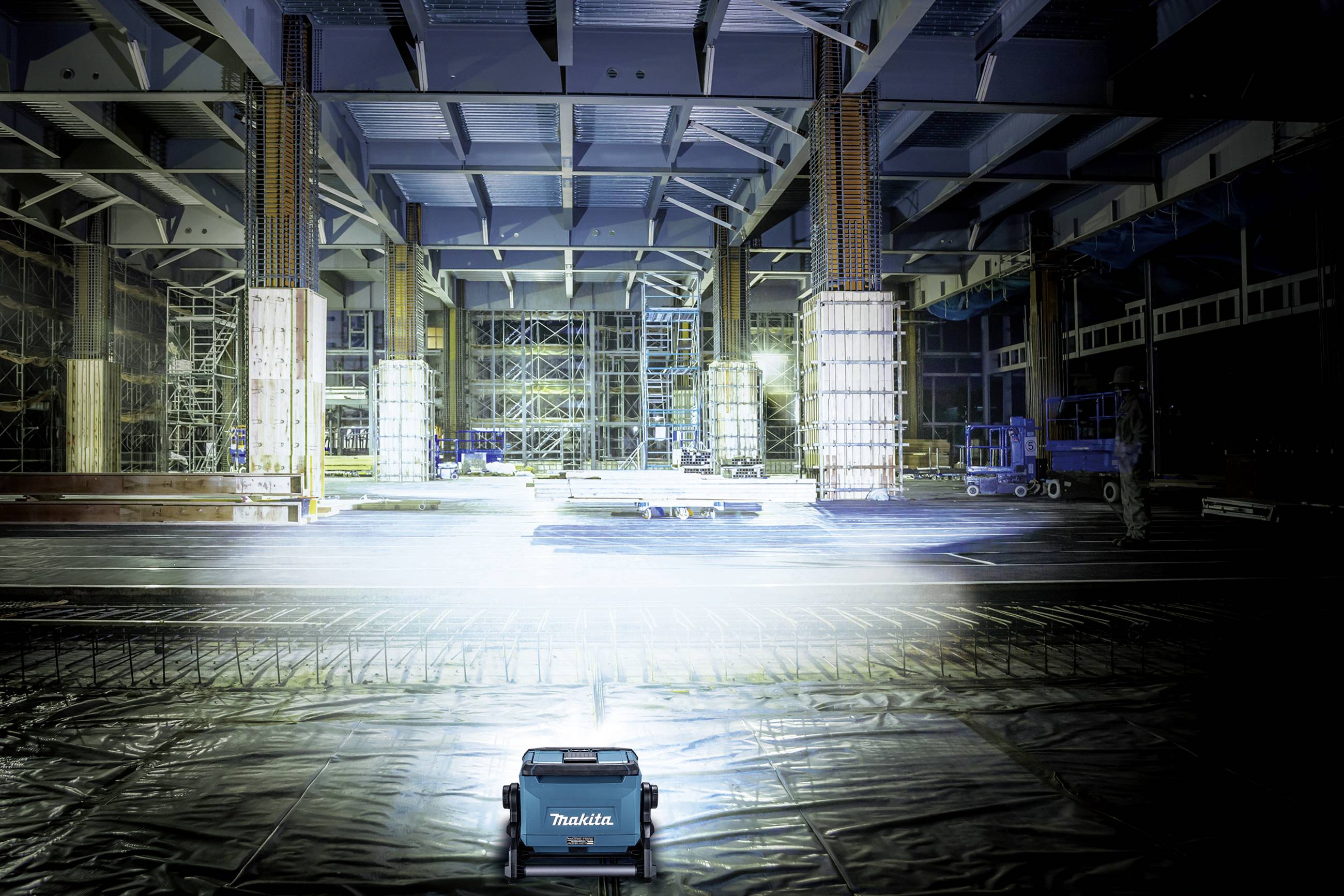Un grande spazio interno vuoto con soffitti alti e travi d'acciaio, illuminato da lampade da cantiere. In primo piano c'è un piccolo dispositivo blu.