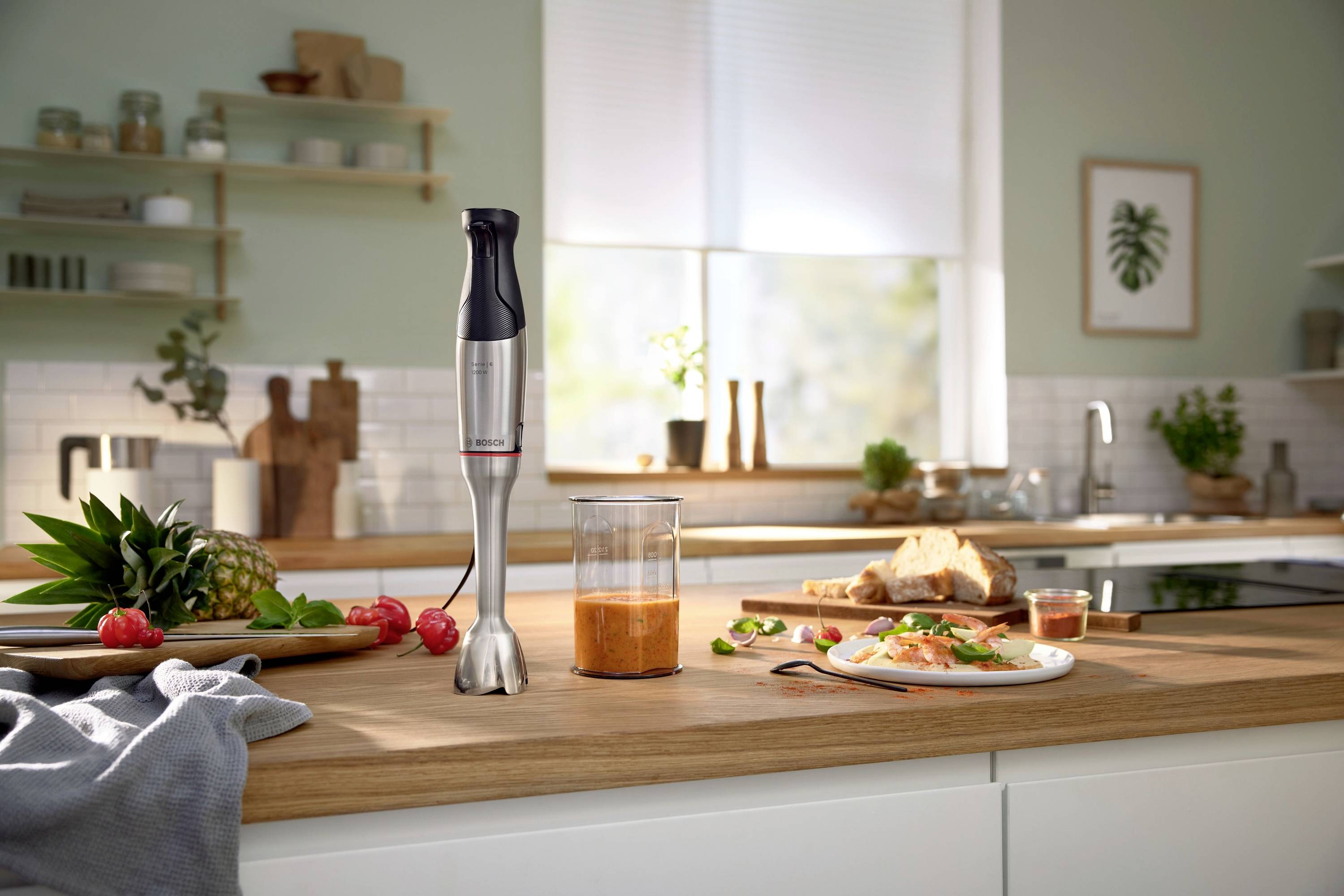 Un frullatore a immersione è posizionato su un piano di lavoro da cucina accanto a frutta fresca e un bicchiere di smoothie. Sullo sfondo si trova una cucina moderna.