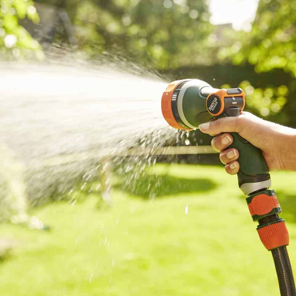 Una mano tiene una pistola a spruzzo da giardino che spruzza acqua in un getto curvo su un prato verde soleggiato.