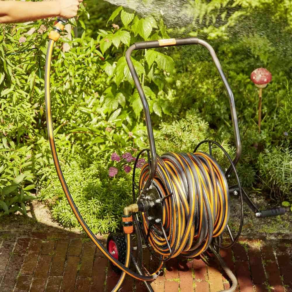 Una persona sta innaffiando un giardino con un tubo dell'acqua avvolto su un avvolgitubo. Sullo sfondo sono visibili delle piante.