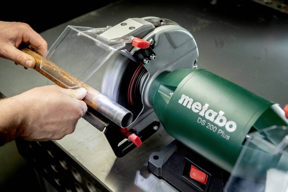 Una persona sta utilizzando una smerigliatrice Metabo DS 200 Plus per rimuovere la ruggine da un tubo metallico. Le mani tengono il tubo in modo stabile.