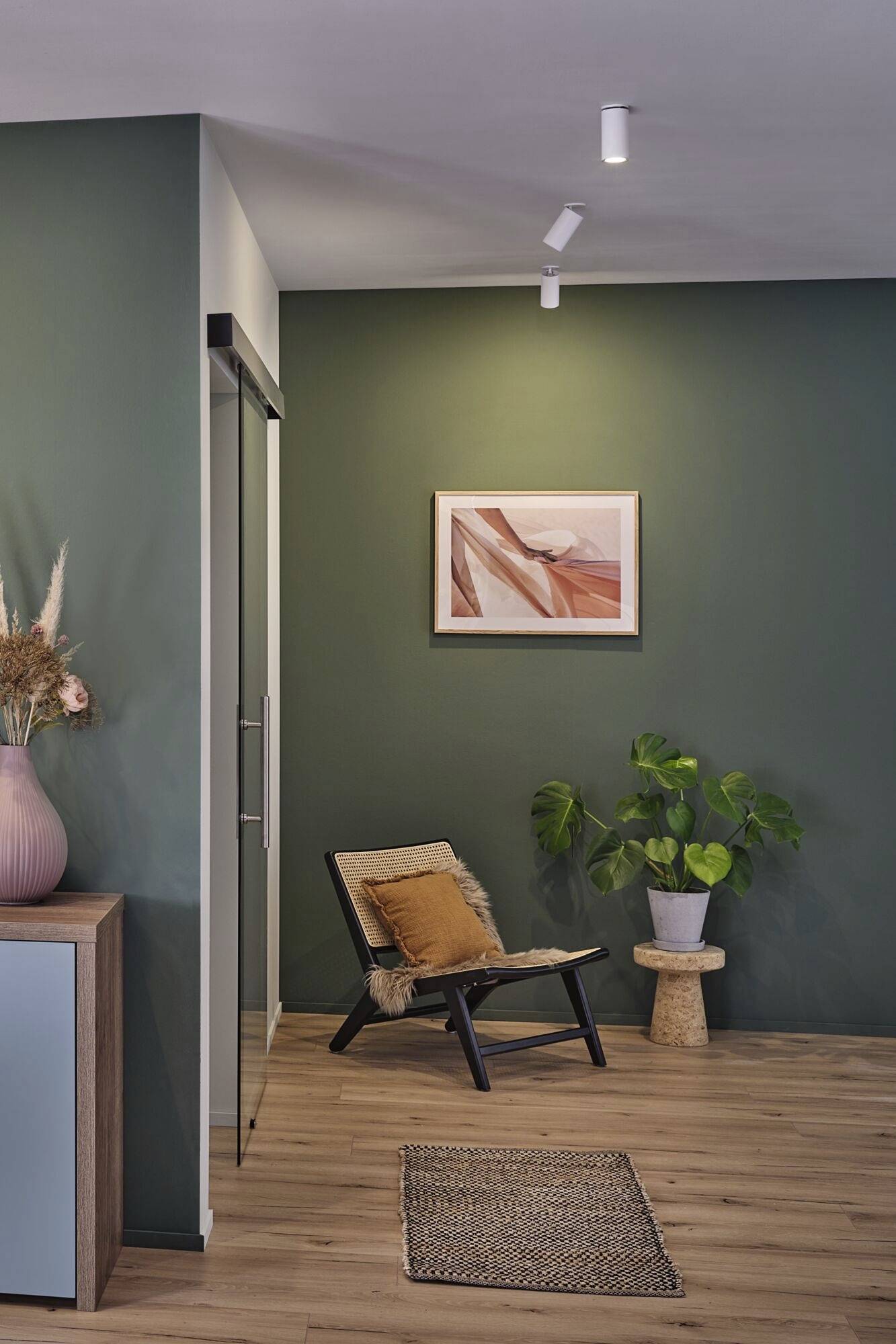 Un soggiorno con una parete dipinta di verde, decorata con un quadro incorniciato. Una sedia con un cuscino è posizionata accanto a una pianta.